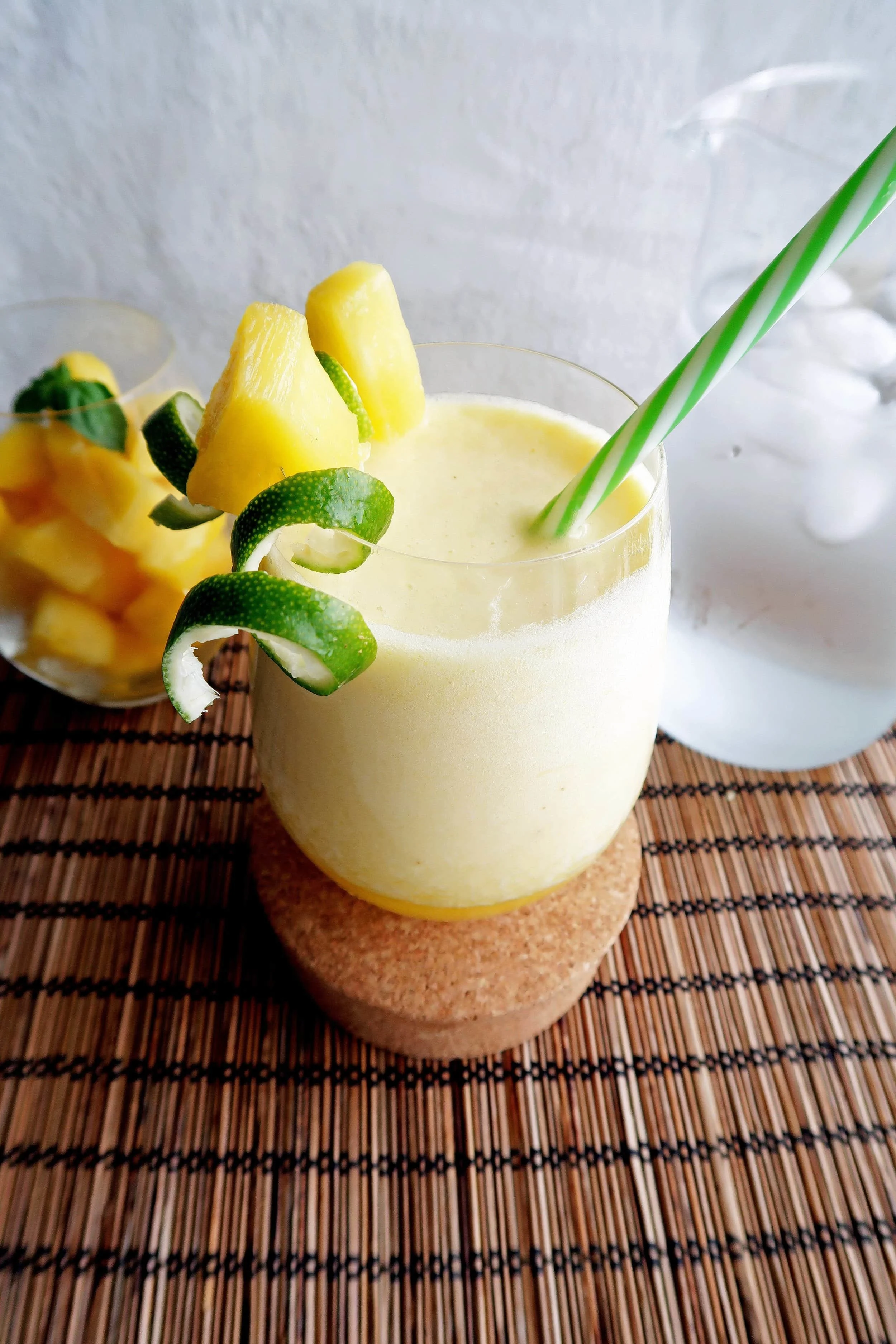 A glass of Pineapple Coconut Water Slushie with lime rind and pineapple garnish with glasses of pineapples chunks and water in the background.