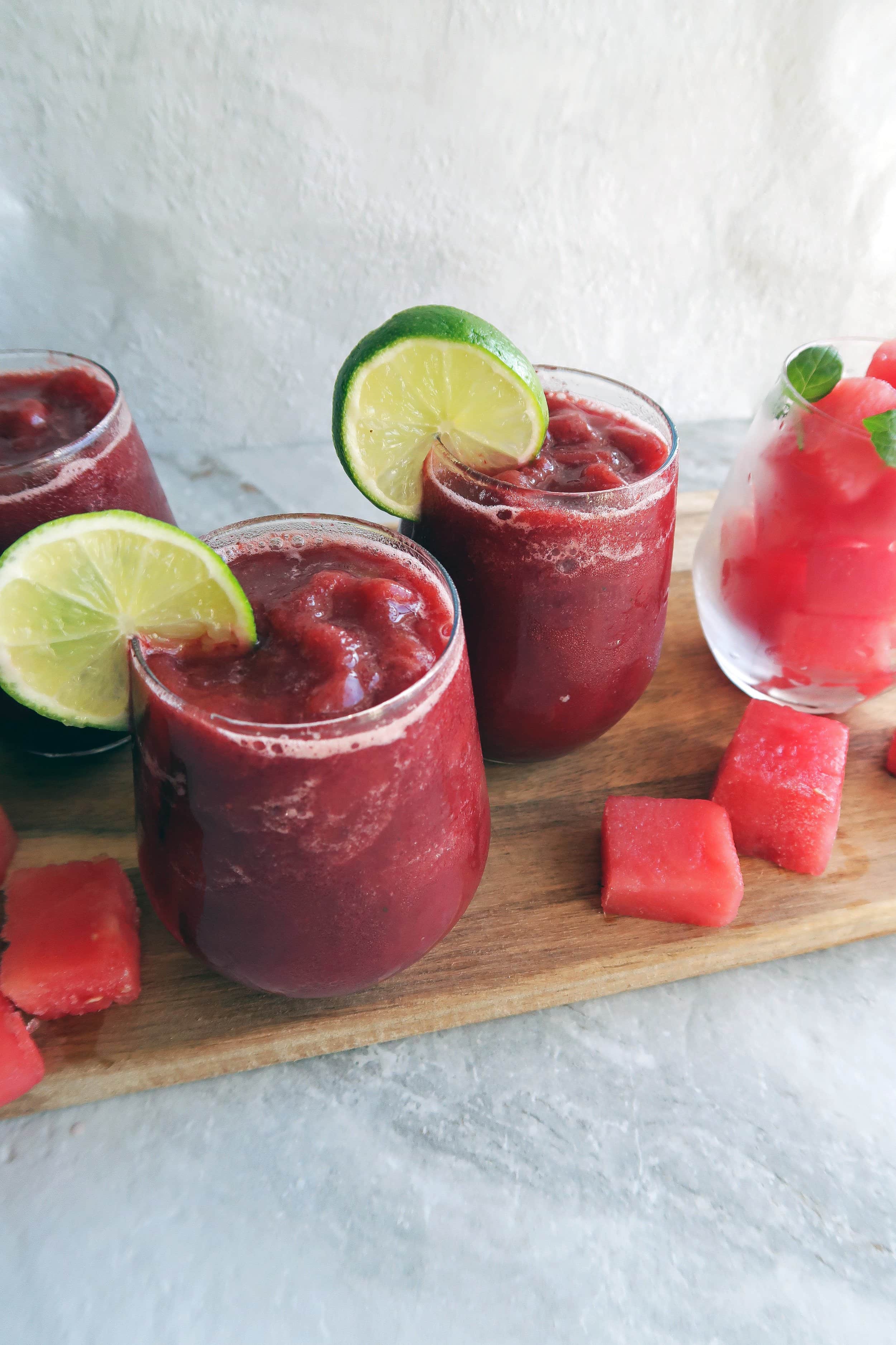 Three Watermelon Cherry Slushies in round glass cups on a wooden board with watermelon cubes around it.