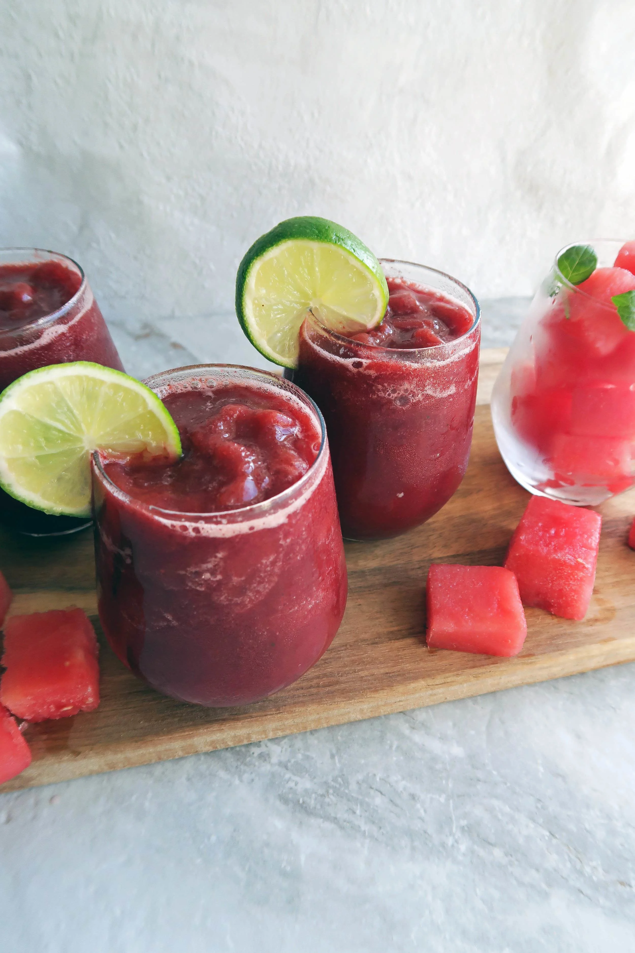 Three Watermelon Cherry Slushies in round glass cups on a wooden board with watermelon cubes around it.