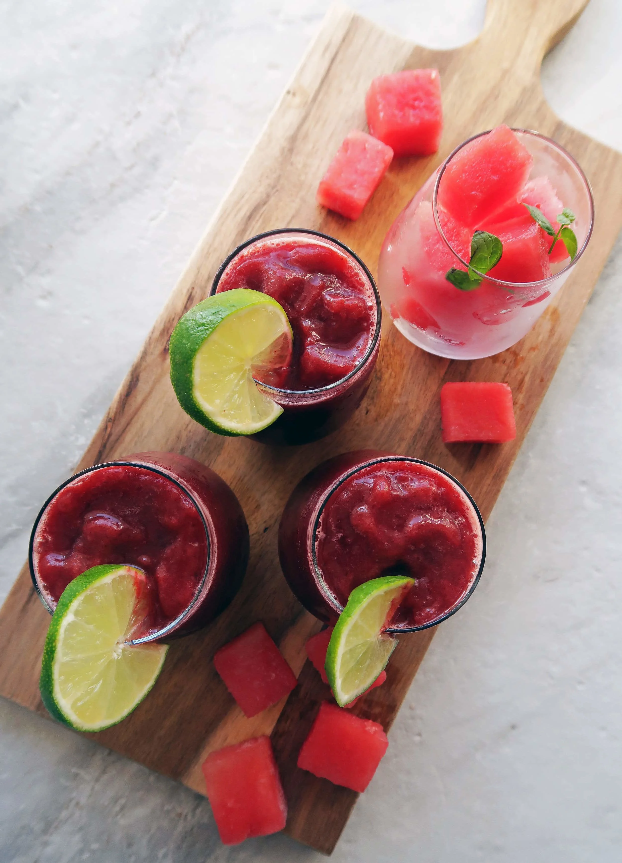 Overhead view of three Watermelon Cherry Slushies in glass cups with a lime slice on each rim, watermelon cubes around it, another glass full of watermelon pieces.