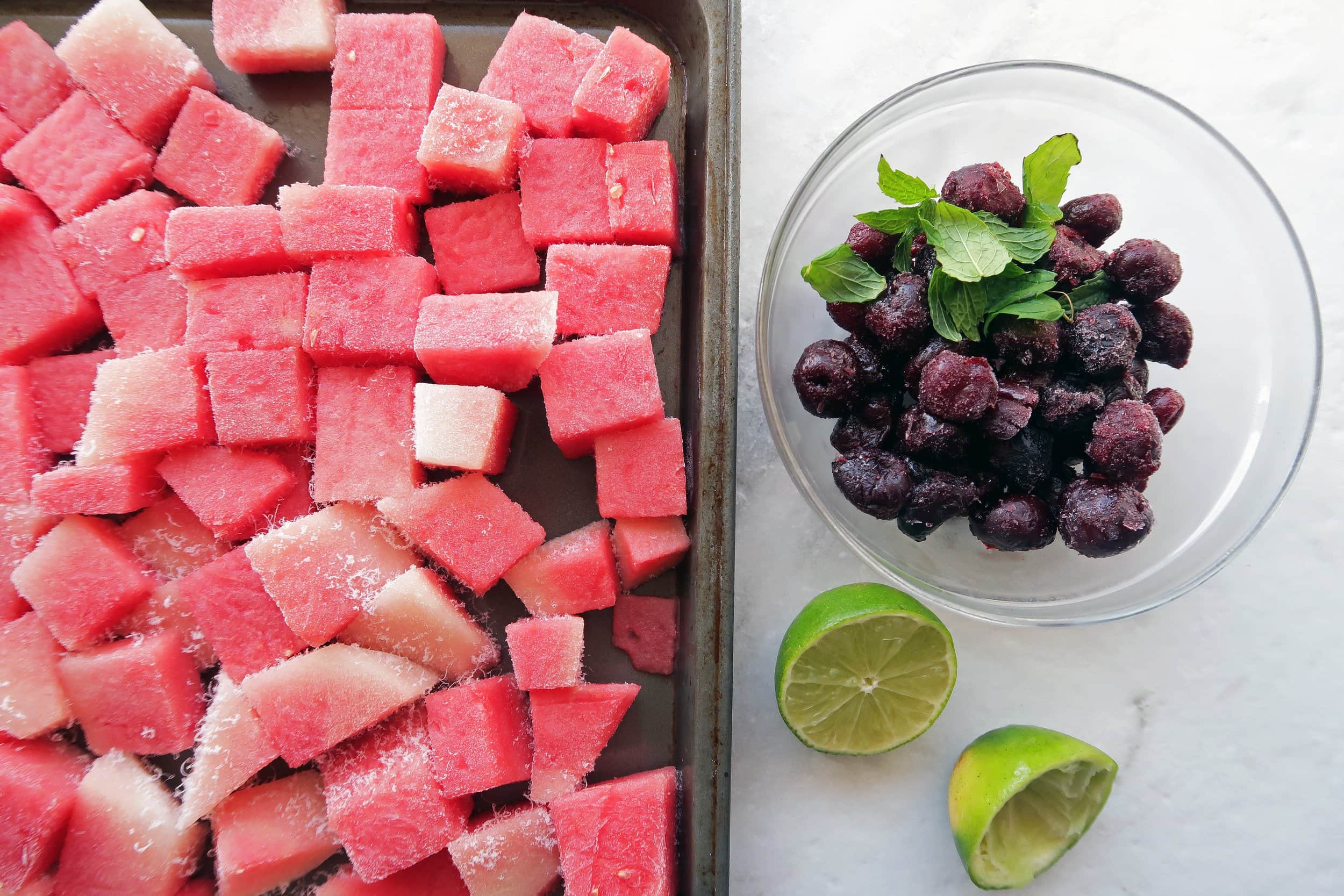 Frozen cubed watermelon on a baking sheet, frozen cherries in a bowl, and a lime on the side.