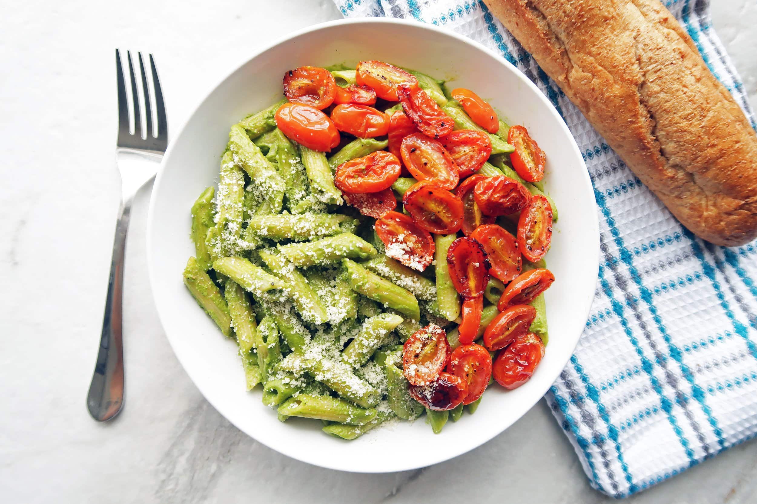 Pesto pasta with roasted tomatoes in a white bowl; a fork, kitchen towel, and french bread to its side.