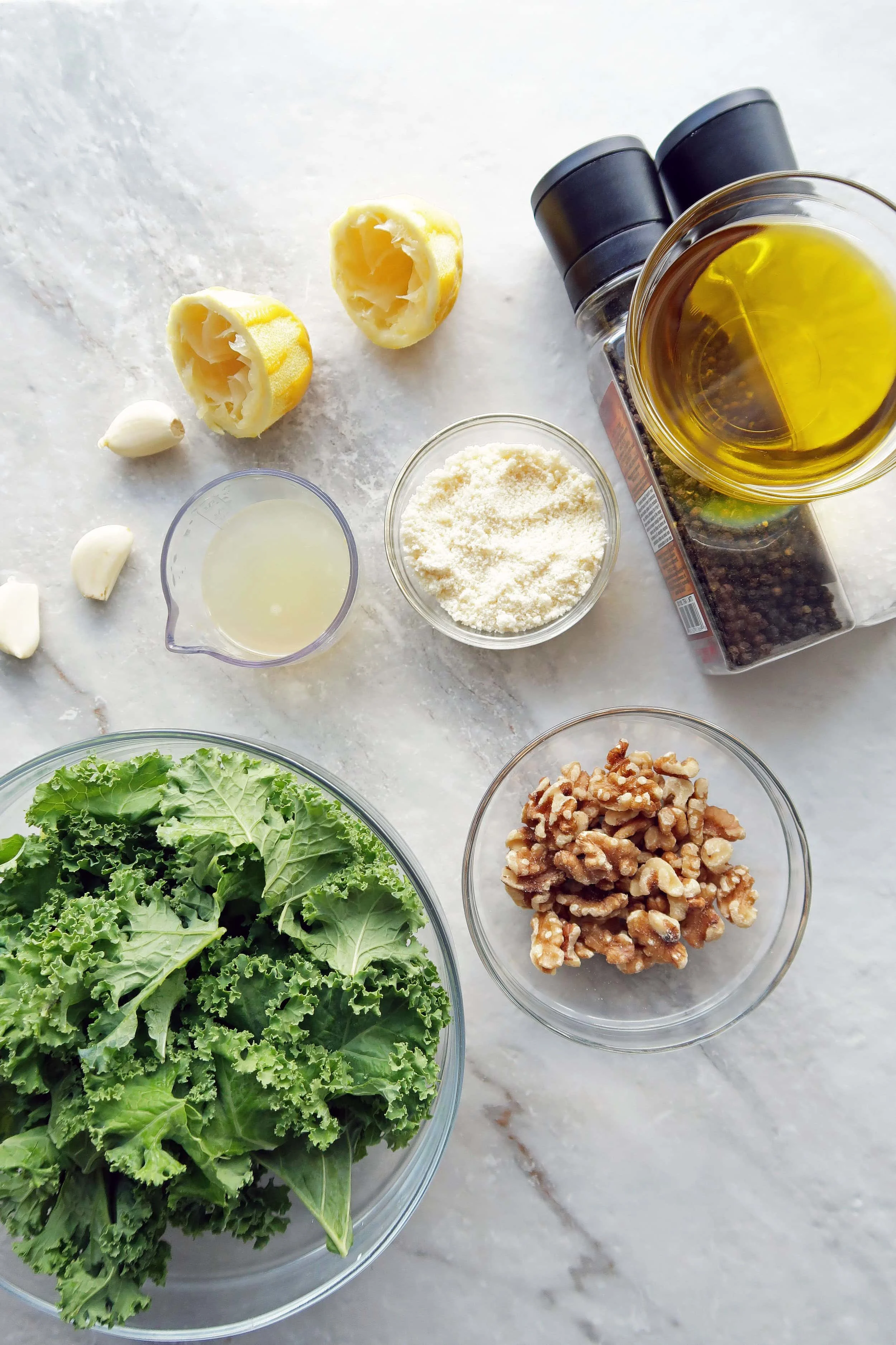 Bowls of kale, walnuts, olive oil, lemon juice, and parmesan cheese along with loose garlic and salt and pepper shakers.