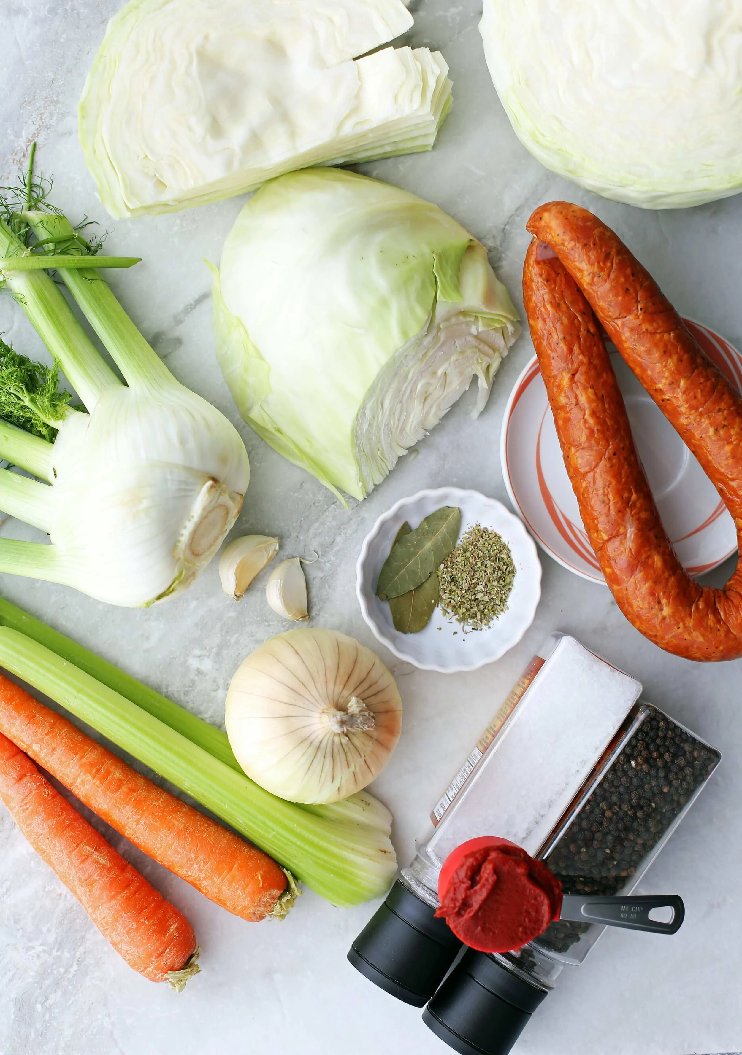 Carrots, celery, onion, garlic, cabbage, fennel, Kielbasa sausage, dried oregano, bay leaf, tomato paste, and salt and pepper on a marble counter.