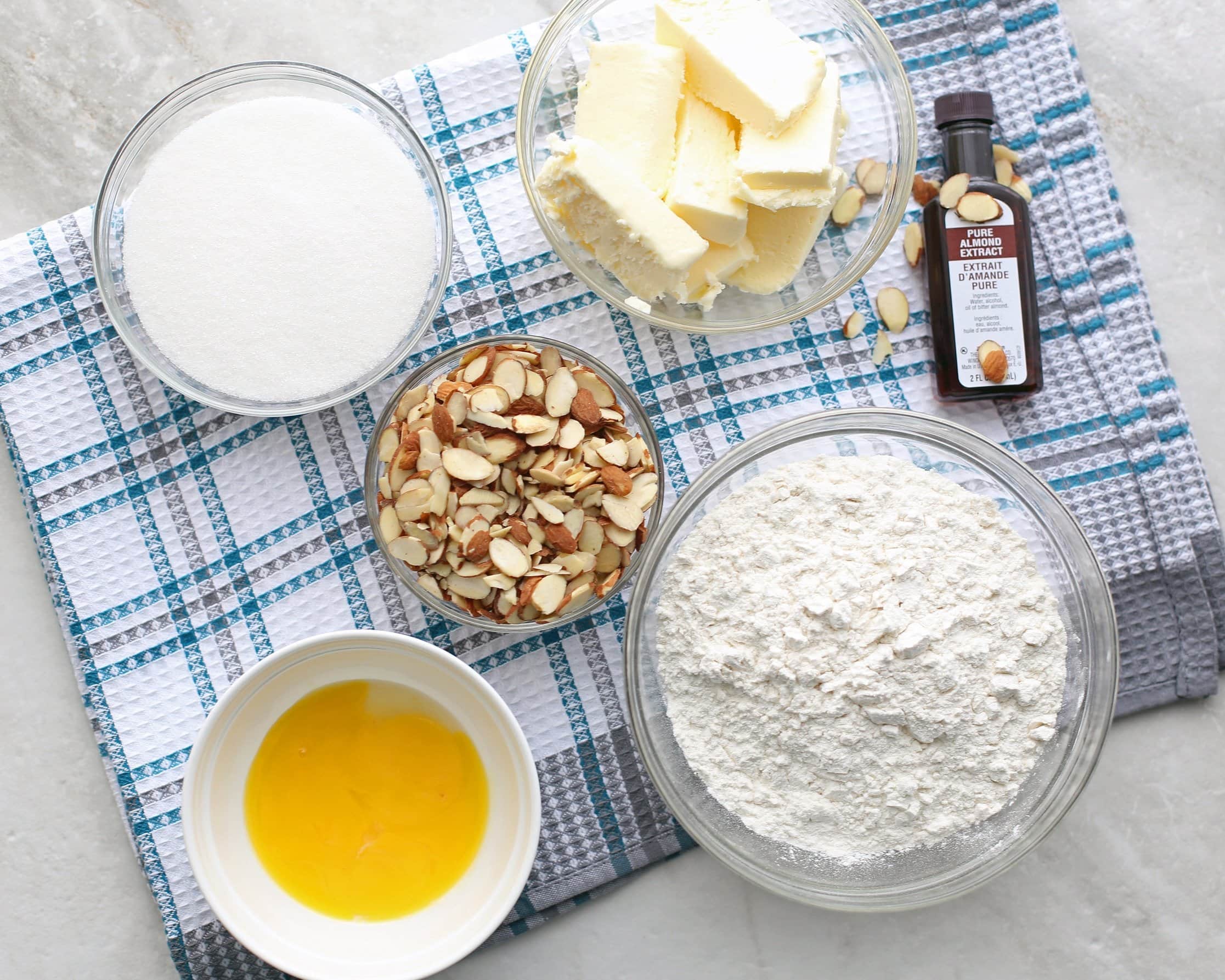 Overhead view of flour, sugar, butter, egg yorks, vanilla extract, and sliced almonds.