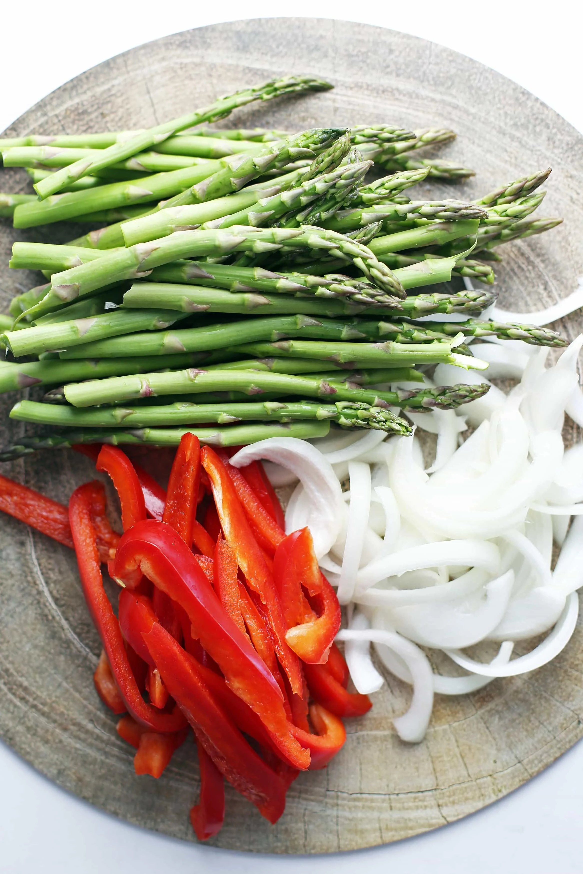 Sliced red bell pepper, sliced white onion, and trimmed asparagus on a round wooden platter.