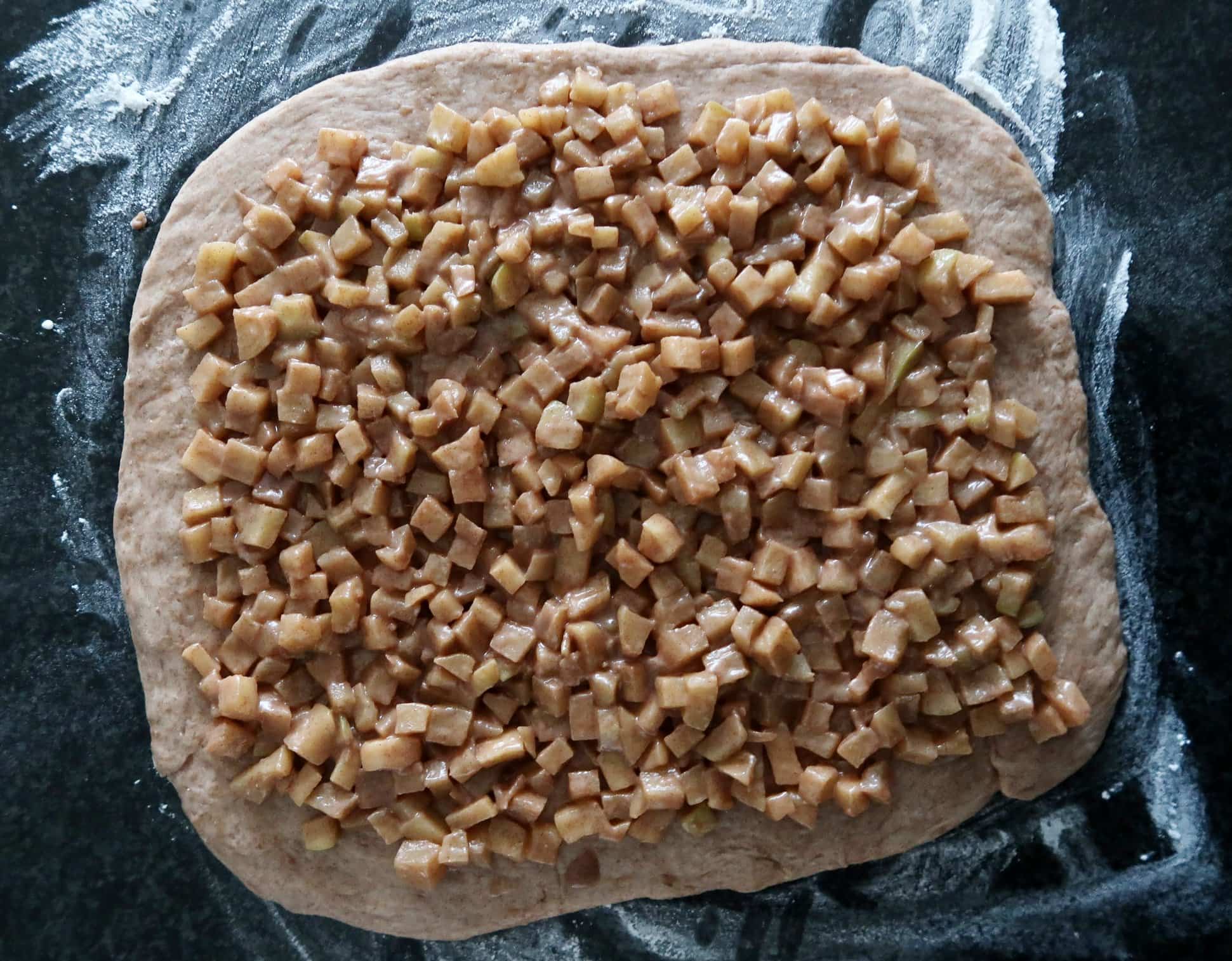 Cinnamon roll dough rolled out on a floured surface and topped with apple filling.