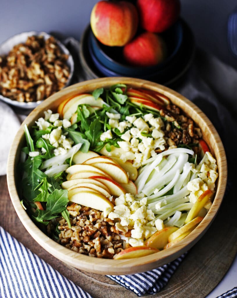 A large bowl of apple fennel arugula salad, a small bowl of walnuts, and three gala apples in a blue bowl.