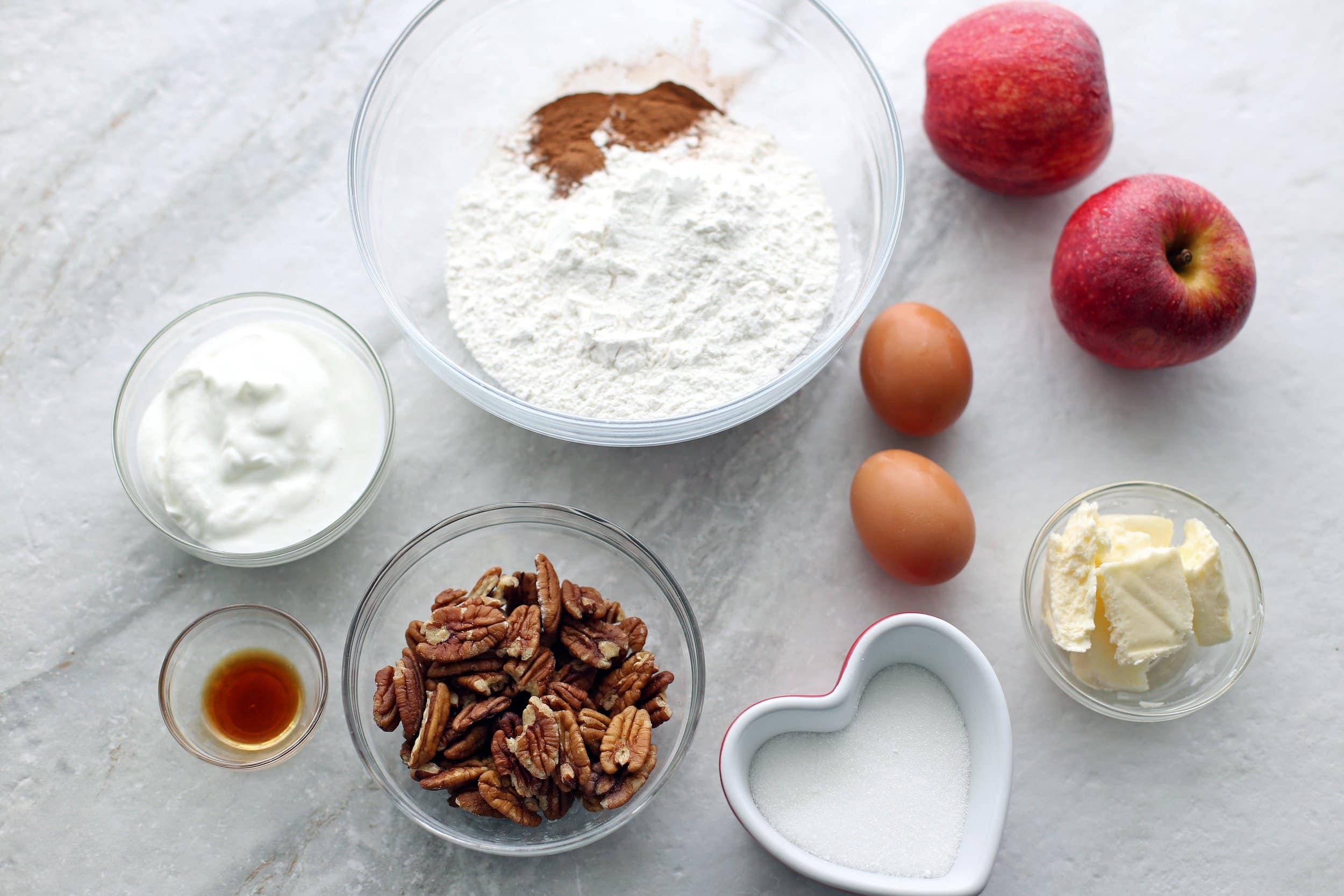 Overhead view of flour mixture, cinnamon, greek yogurt, vanilla extract, pecans, eggs, butter, and apples.