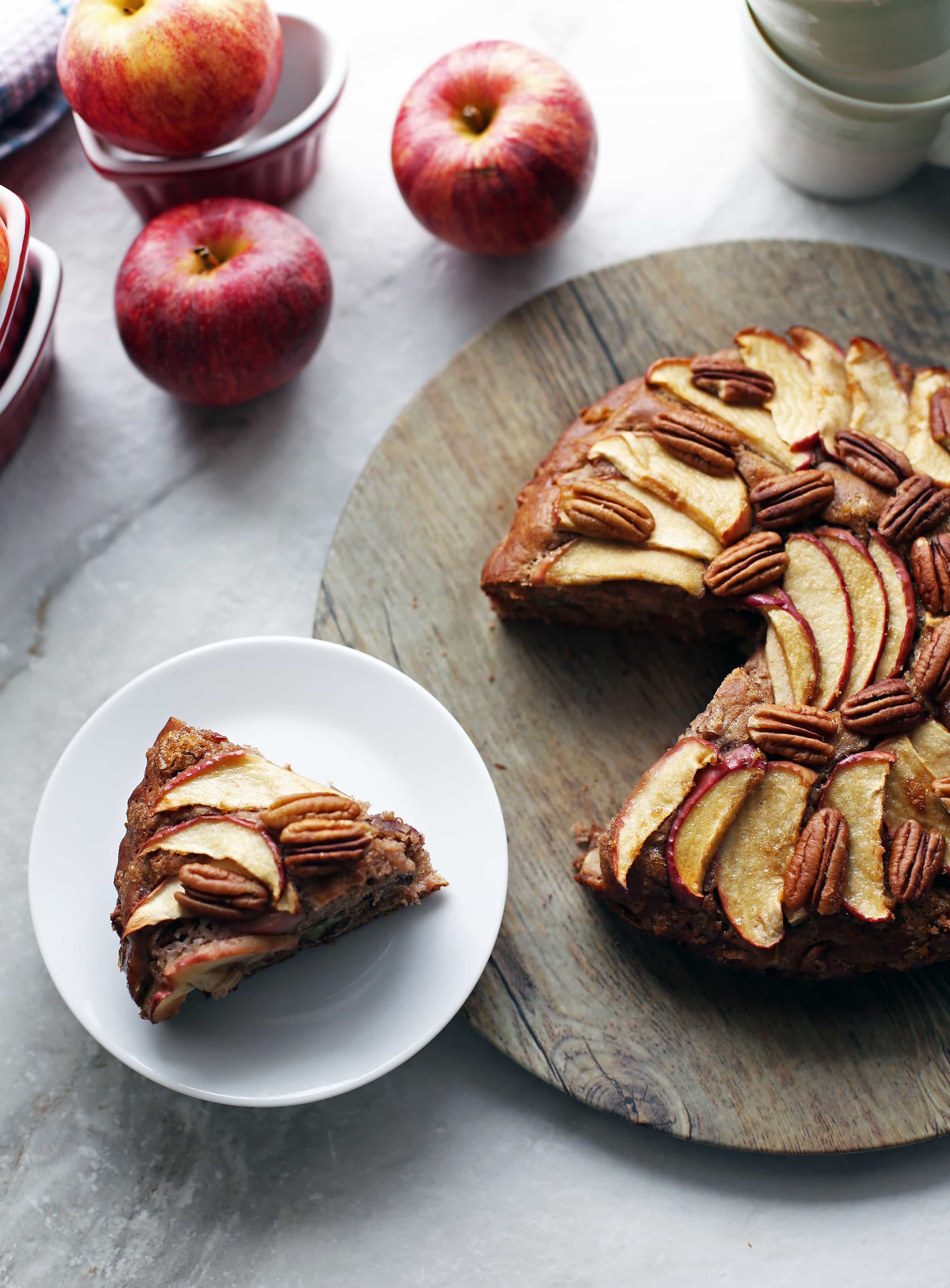 Rustic apple cinnamon pecan cake on a round wooden platter with a single slice on a white plate.