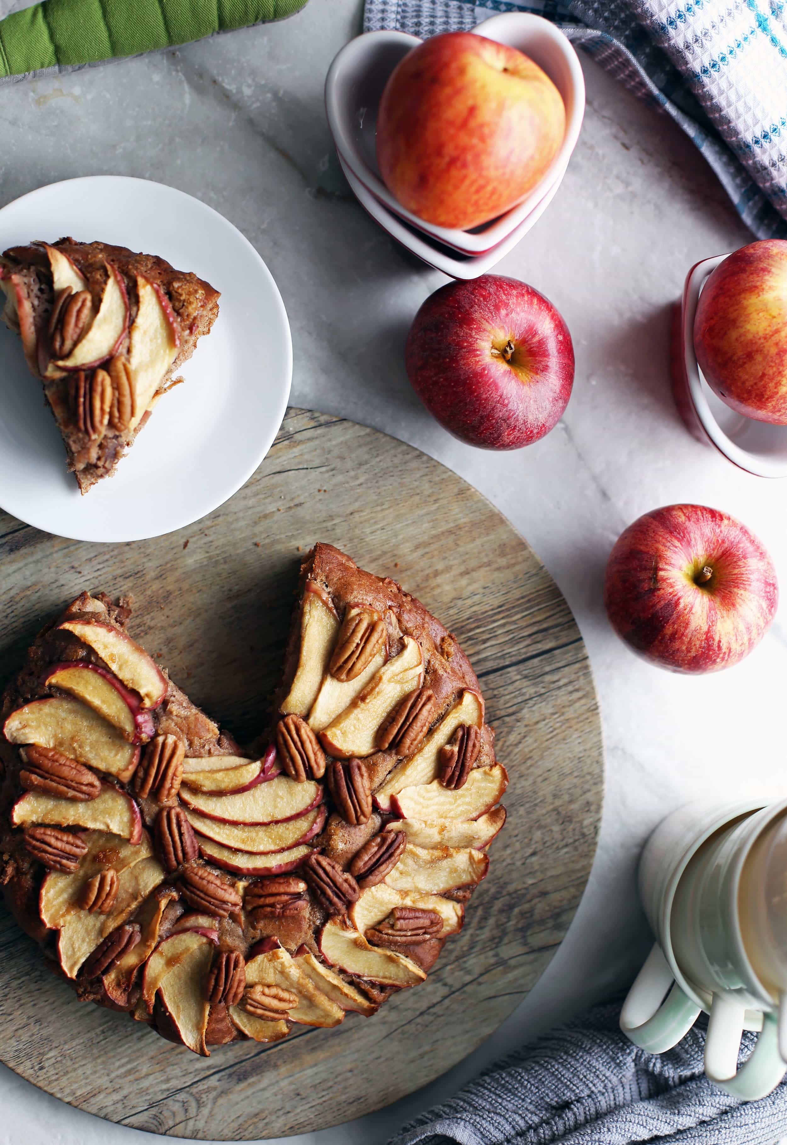 Rustic apple cinnamon pecan cake on a round wooden platter with a single slice on a white plate.