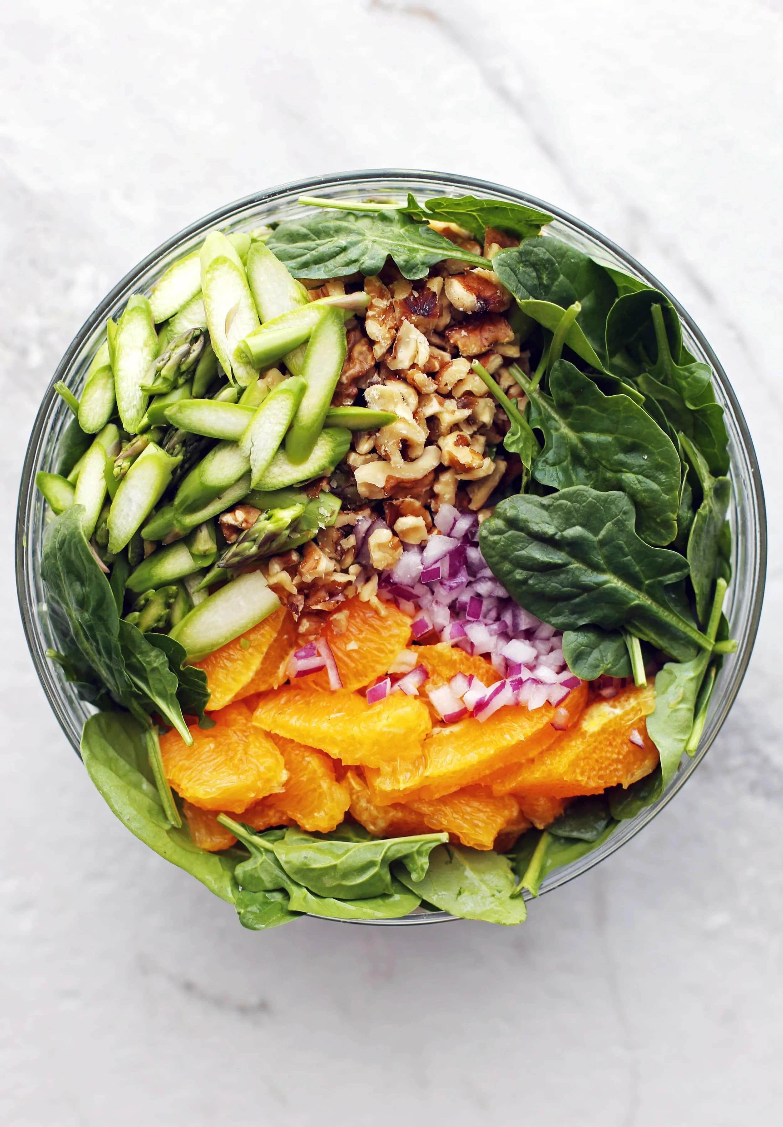 A glass bowl full of asparagus, spinach, orange, red onion, avocado, and walnuts.