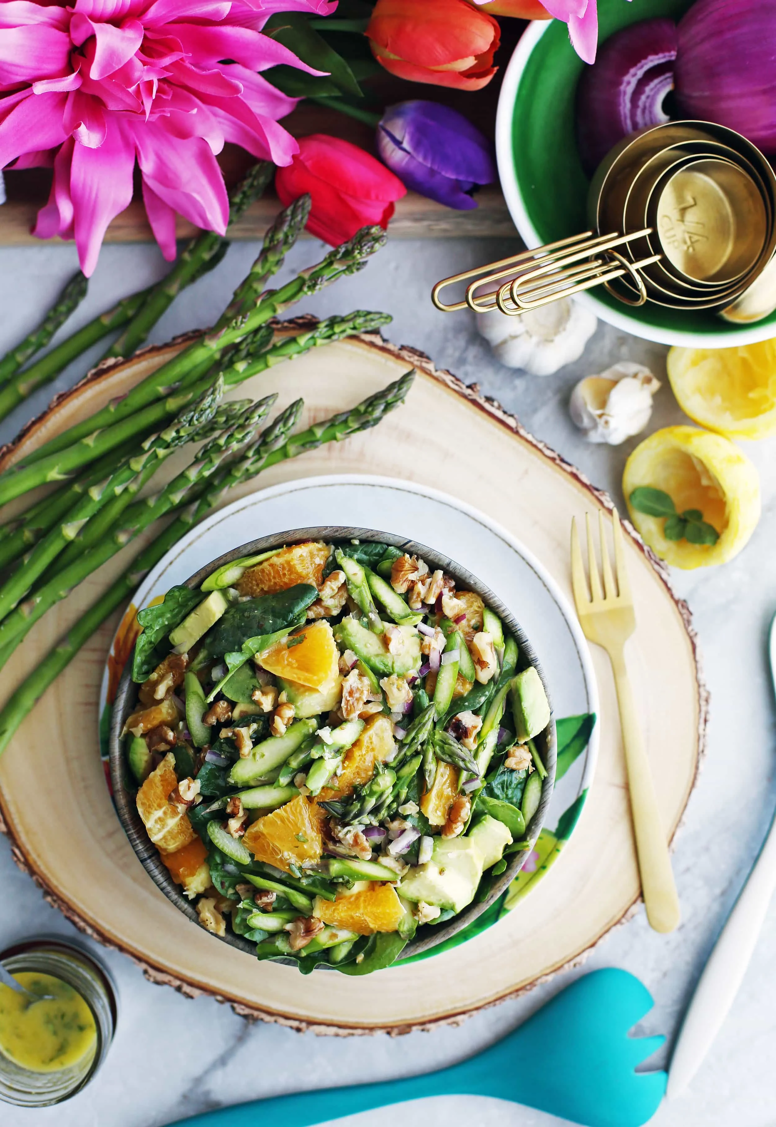 Asparagus Orange Spinach Salad with Basil Lemon Vinaigrette in a wooden bowl with a fork, vinaigrette, and asparagus stalks around it.