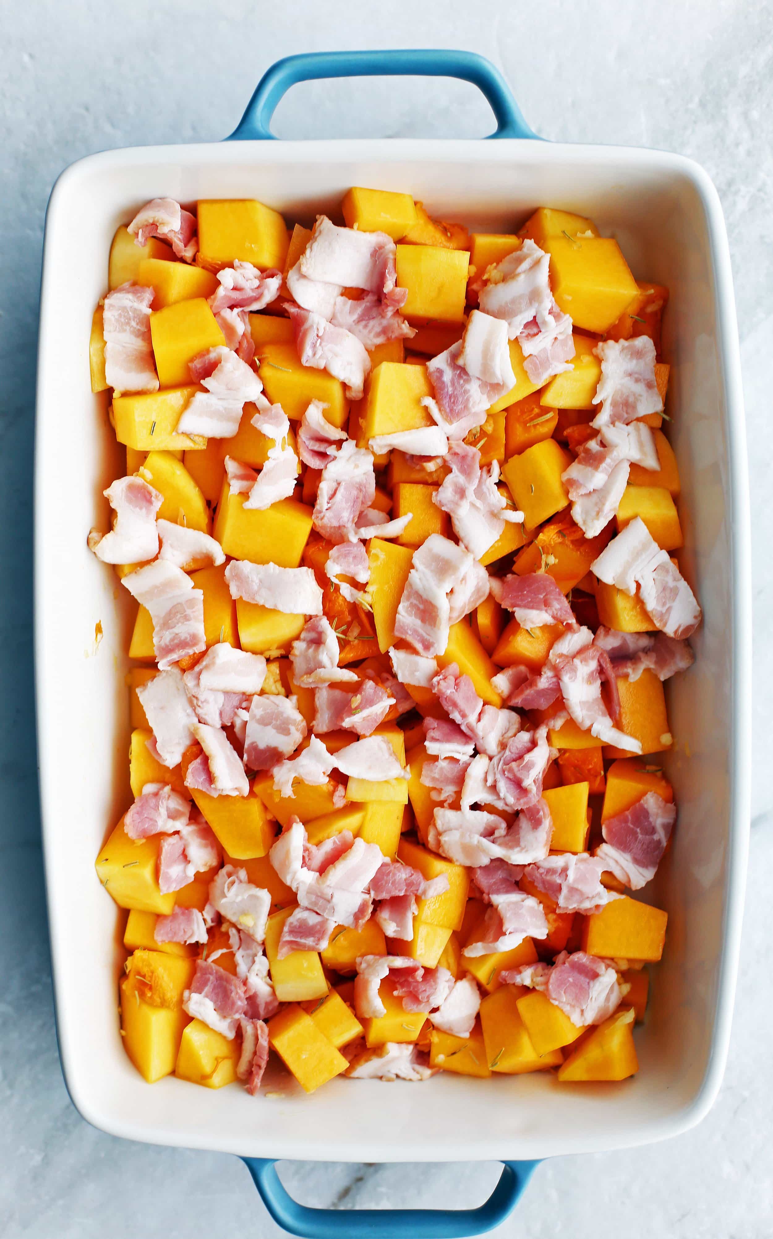 Seasoned chopped butternut squash in a baking dish that’s topped with chopped uncooked bacon.