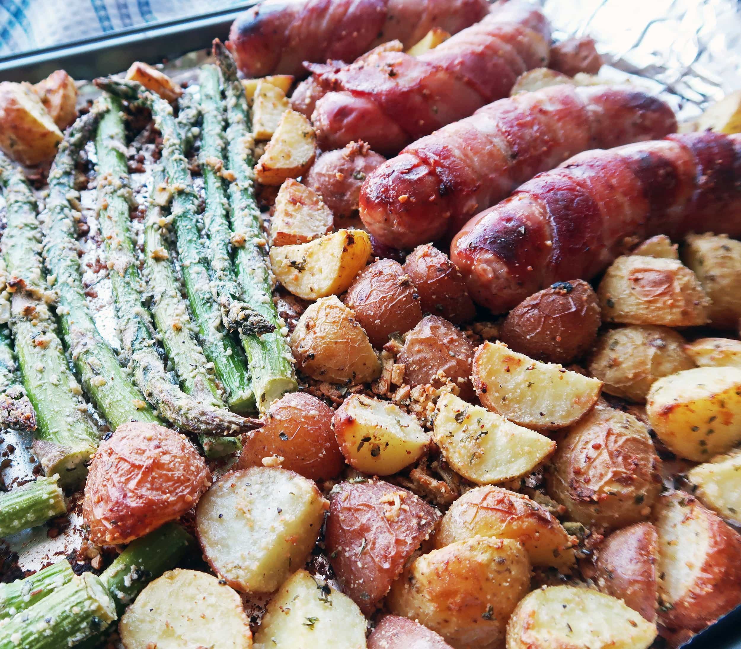 A close-up of parmesan covered potatoes and asparagus with bacon-wrapped sausages.