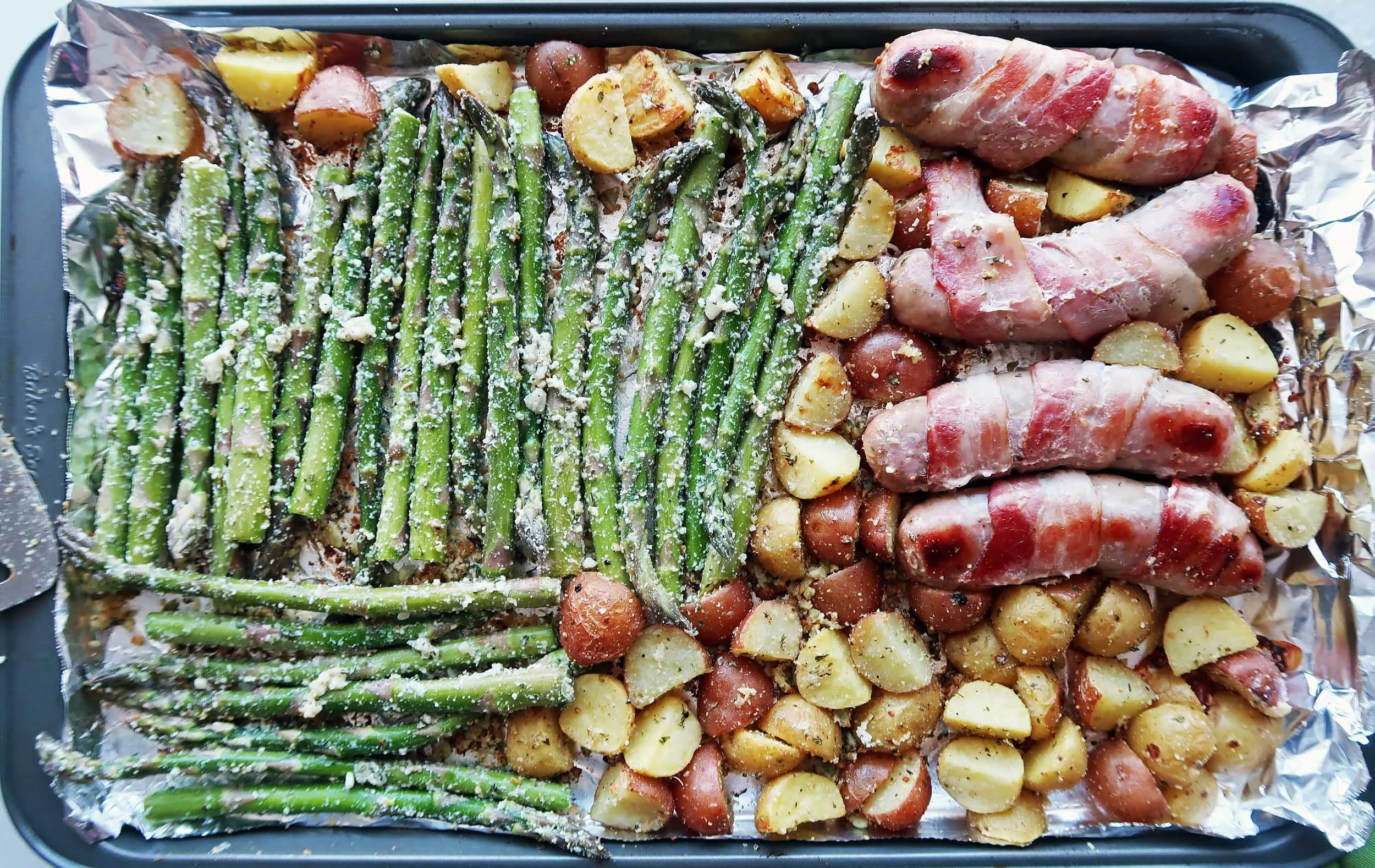 Bacon-wrapped sausages and vegetables on a baking sheet.