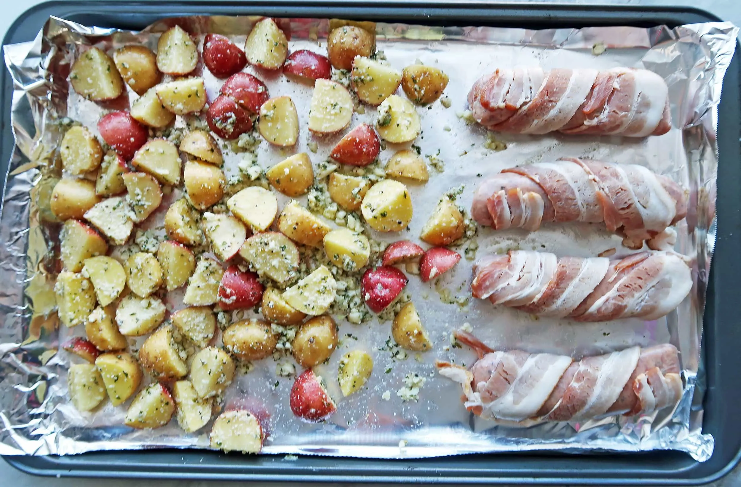 Potatoes and bacon-wrapped sausages on a baking sheet.