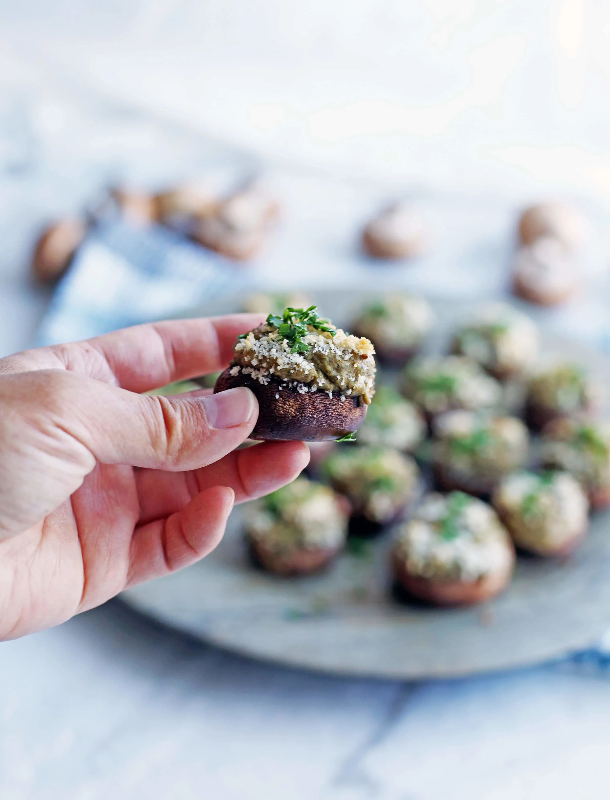 A hand holding a single baked stuffed mushroom on top of a platter containing more stuffed mushrooms.