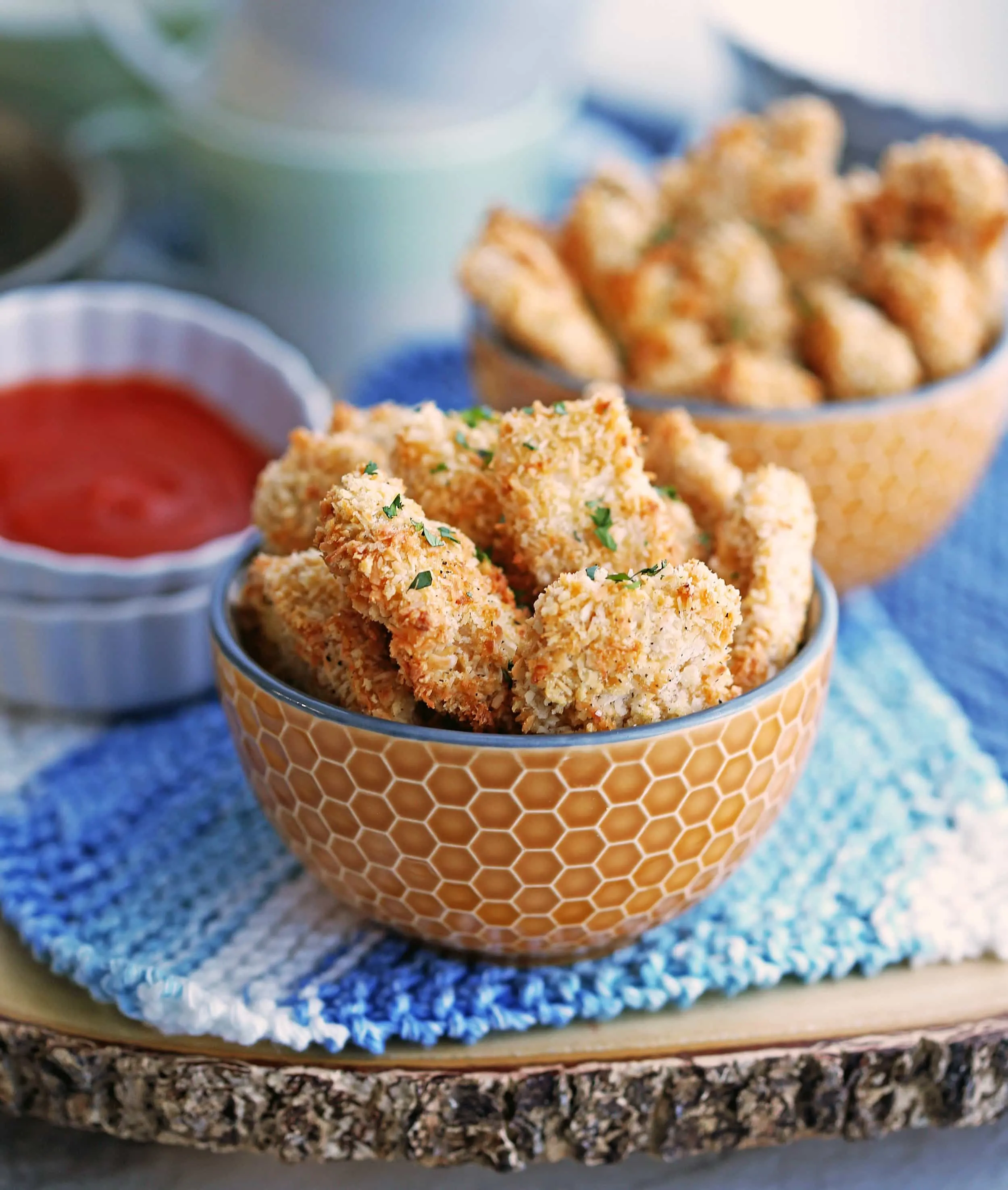 Baked crispy coconut chicken nuggets in an orange bowl with more chicken bites and red sauce in the background.
