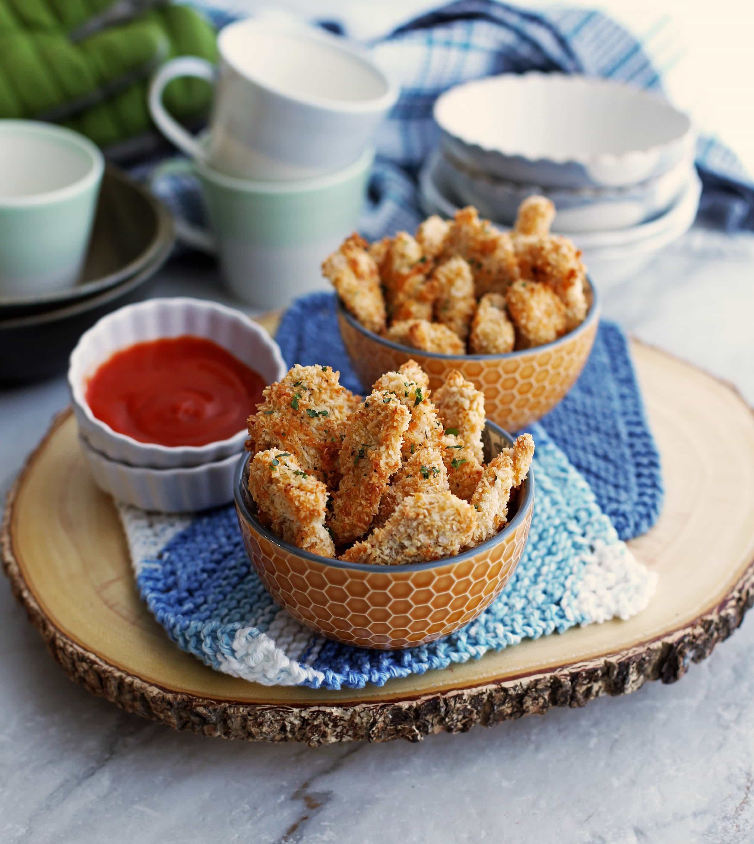 A top angled view of two bowls of baked crispy coconut chicken nuggets with red sauce on the side.