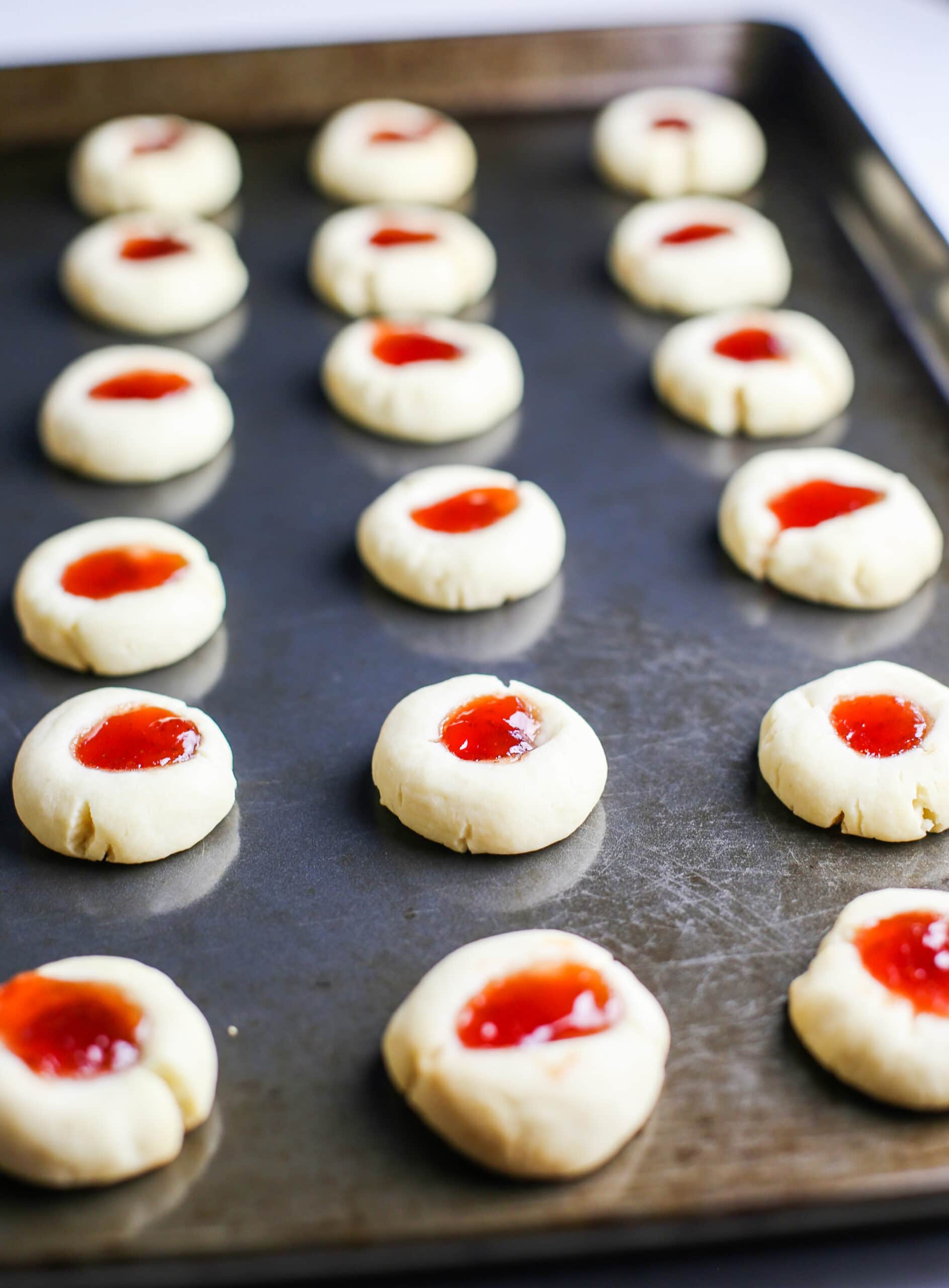 Baked condensed milk thumbprint cookies filled with strawberry jam on a baking sheet.