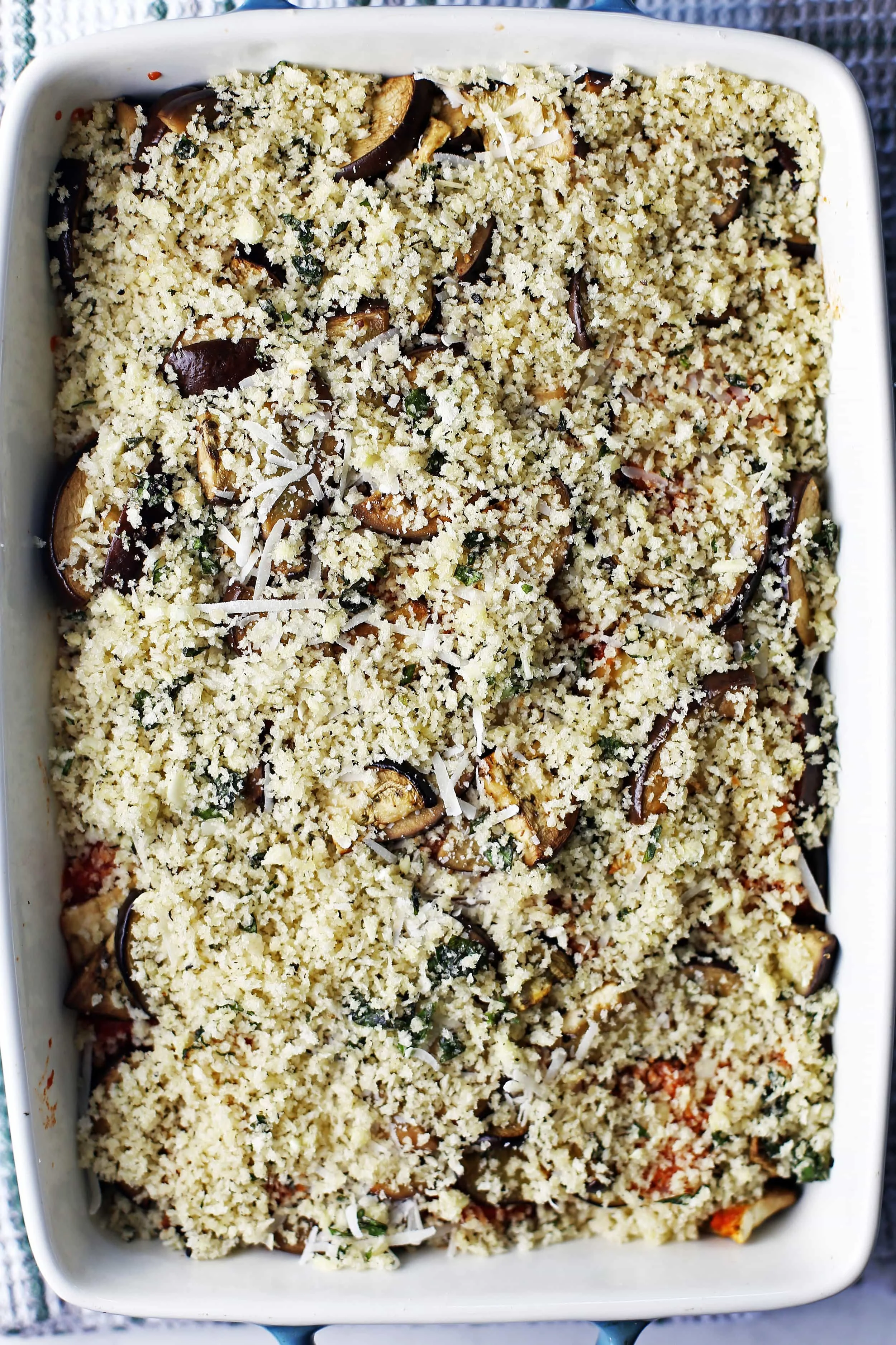 A large casserole dish containing three layers of marinara sauce, sliced eggplant, herbs, and cheeses with a breadcrumb topping.