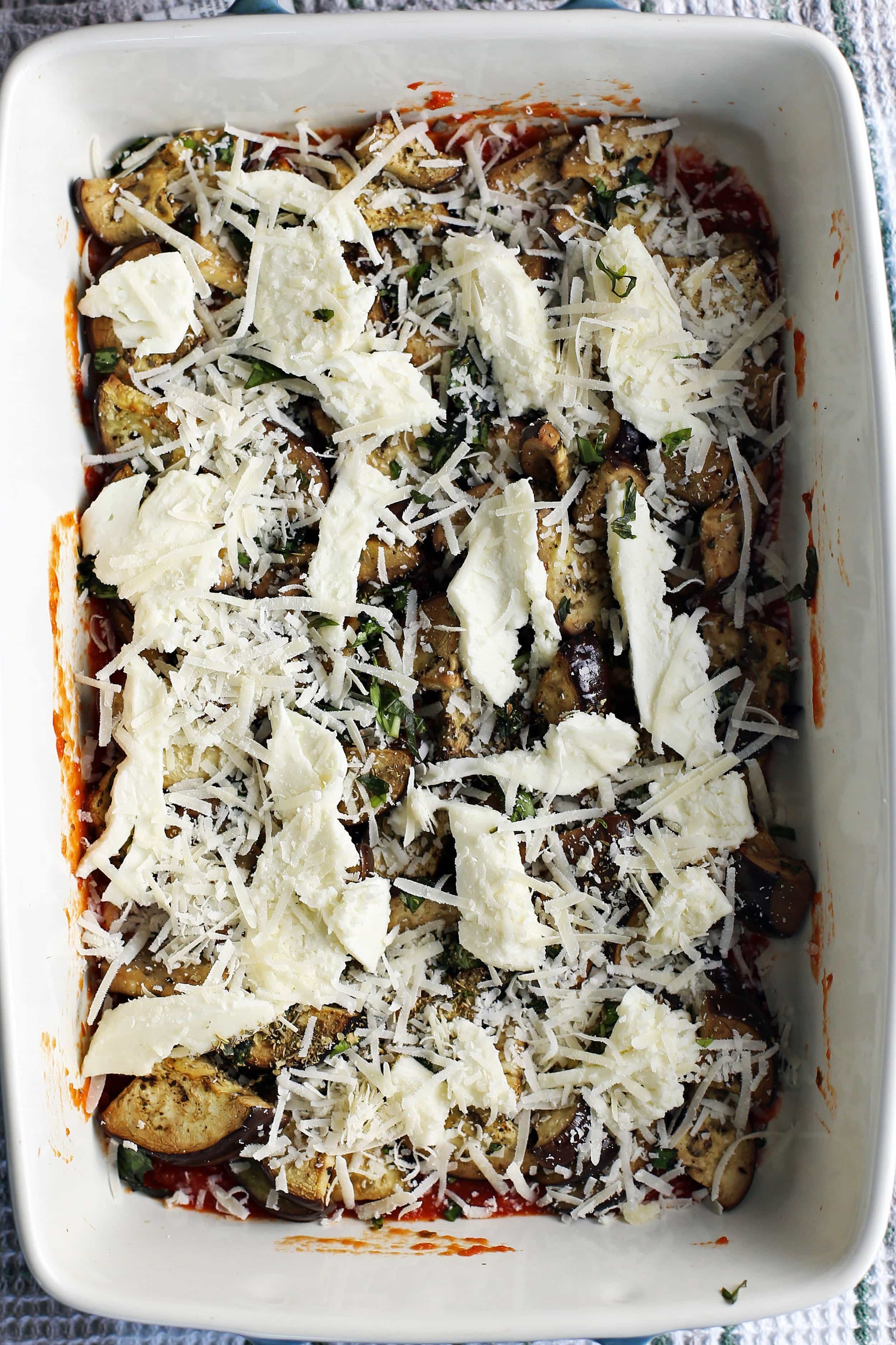 A large casserole dish containing layers of marinara sauce, sliced eggplant, herbs, and cheeses.