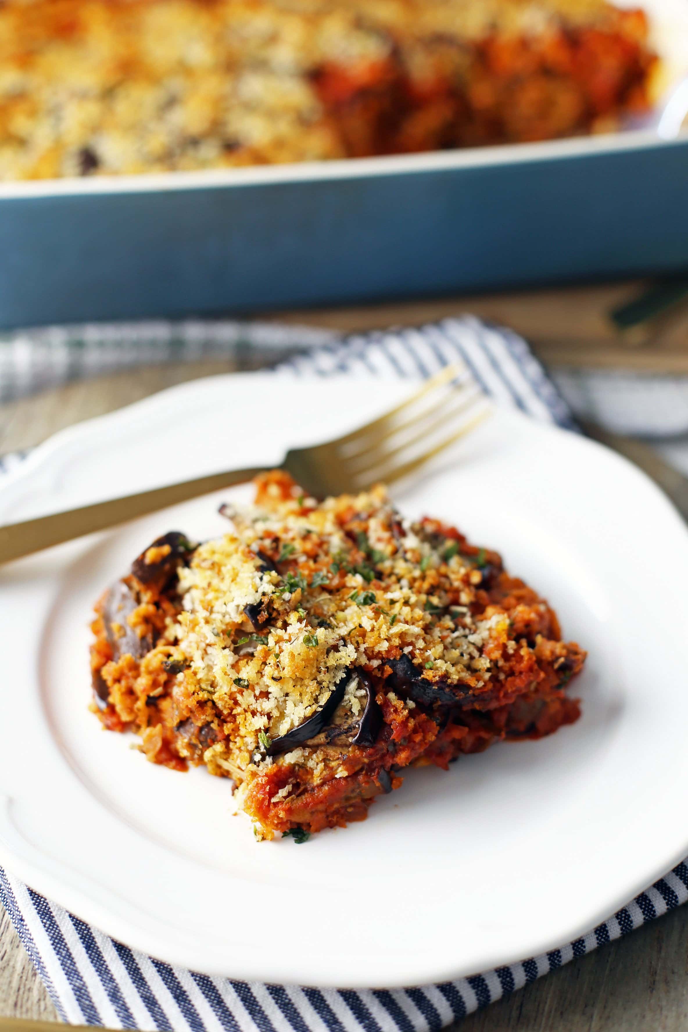 A piece of cheesy layered Baked Eggplant Parmesan on a white plate with a gold fork.