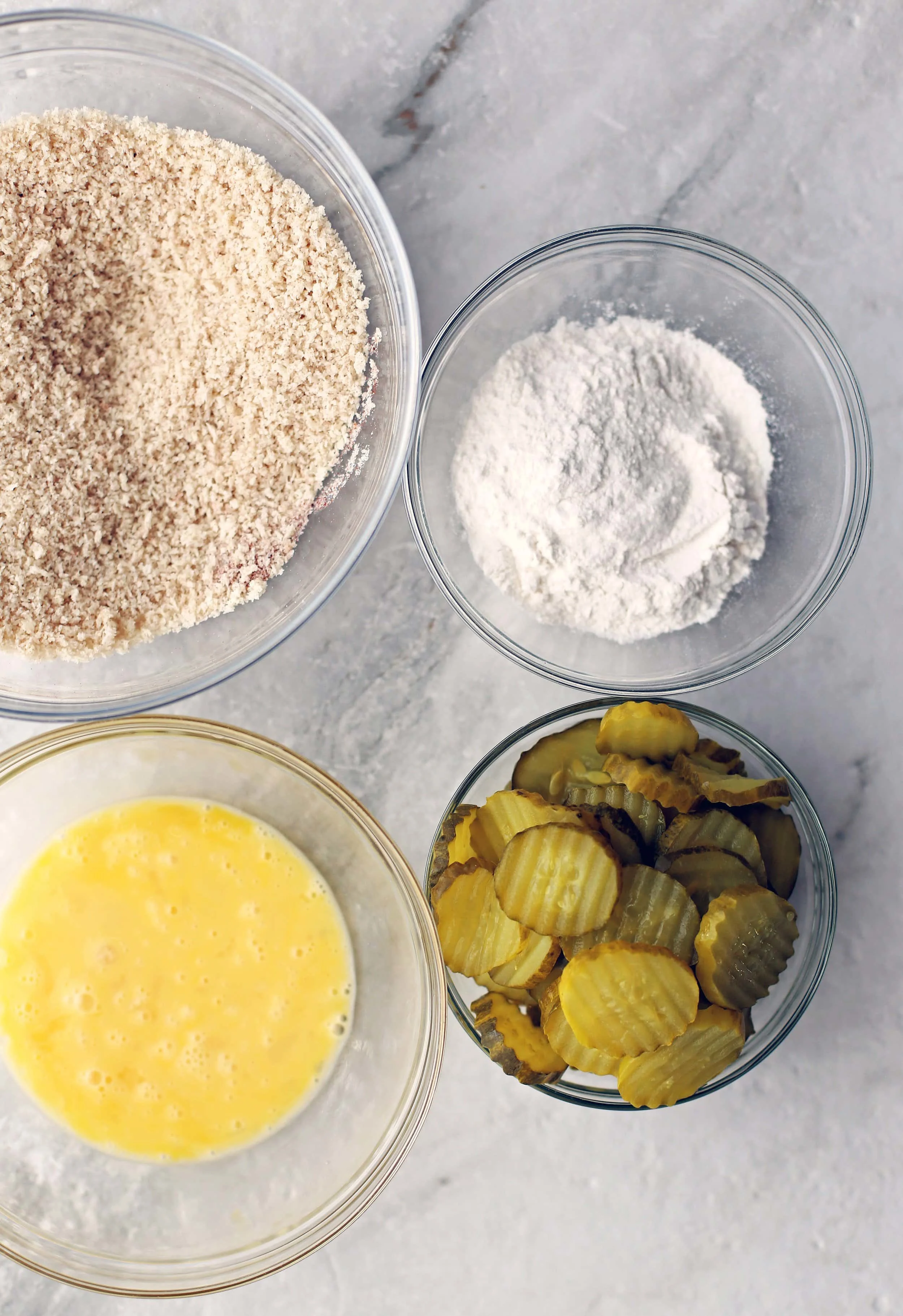 An overhead view of four glass bowls filled with pickle slices, beaten eggs, flour, and panko breadcrumbs.