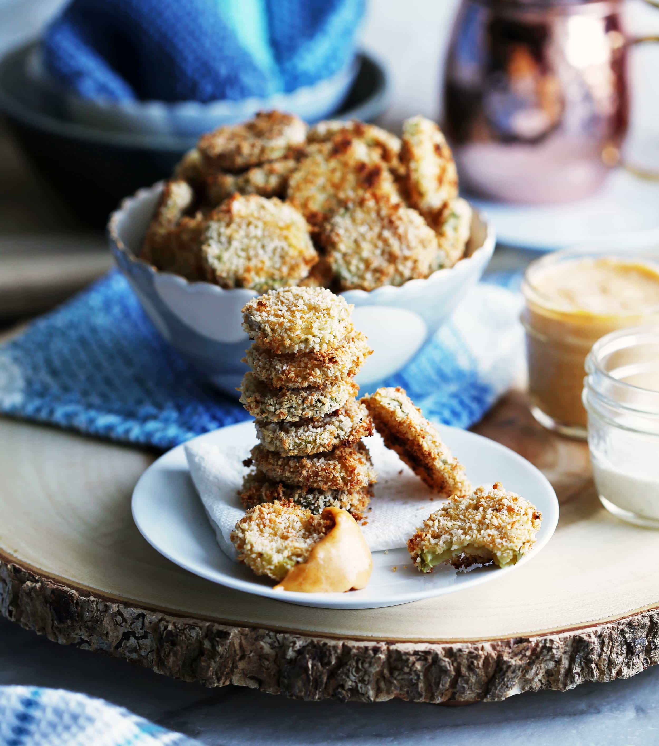 A side view of six stacked crispy oven-fried pickles, one pickle is covered with cheddar sauce, and a bowl of pickles is in the background.