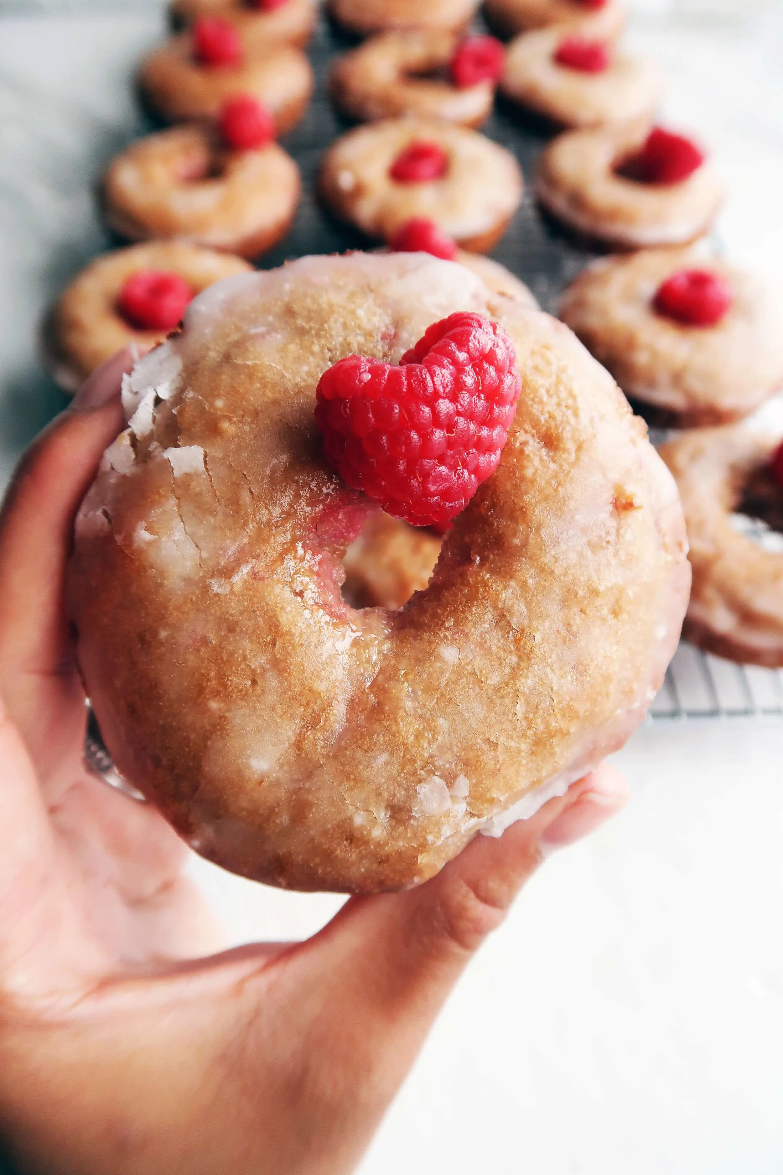 A close-up view of a hand holding a Baked Raspberry Lemon Glazed Donut with more donuts in the background.