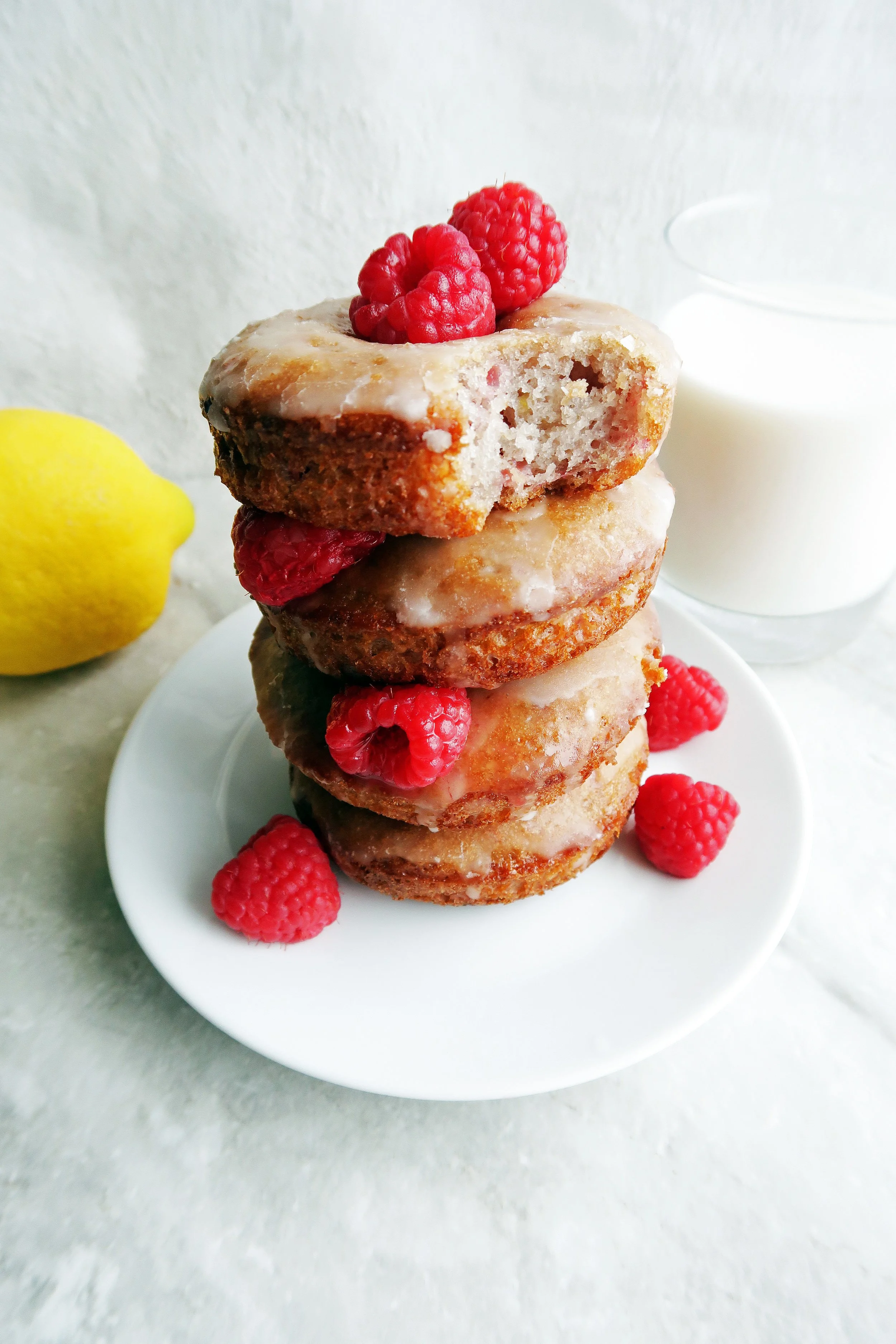 Four Baked Raspberry Lemon Glazed Donuts stacked on a white plate with raspberries around it, a lemon, and glass of milk in the background.