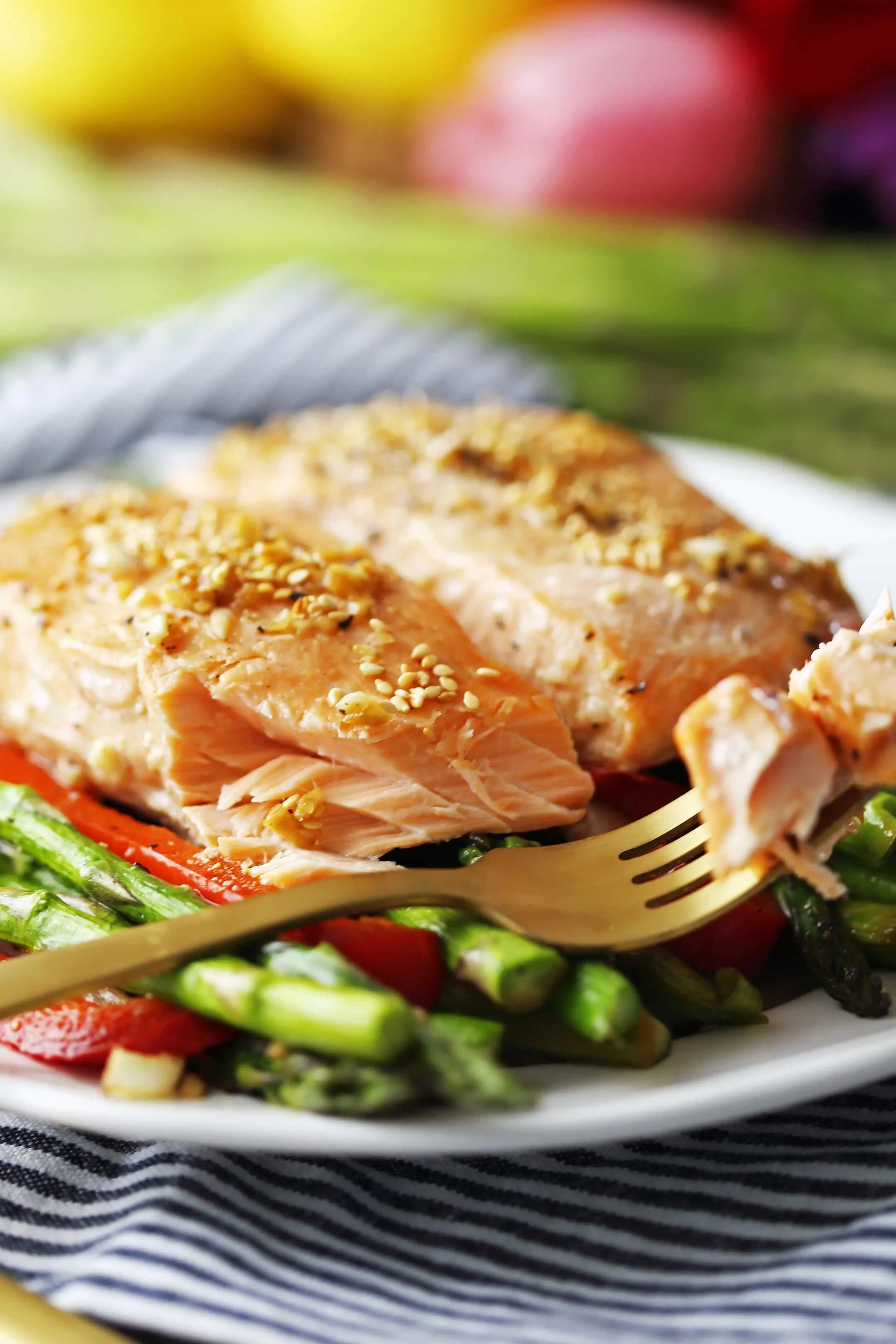 A flaky piece of baked salmon on the tines of a gold fork that’s placed on a white plate with two salmon fillets, asparagus, and bell peppers.
