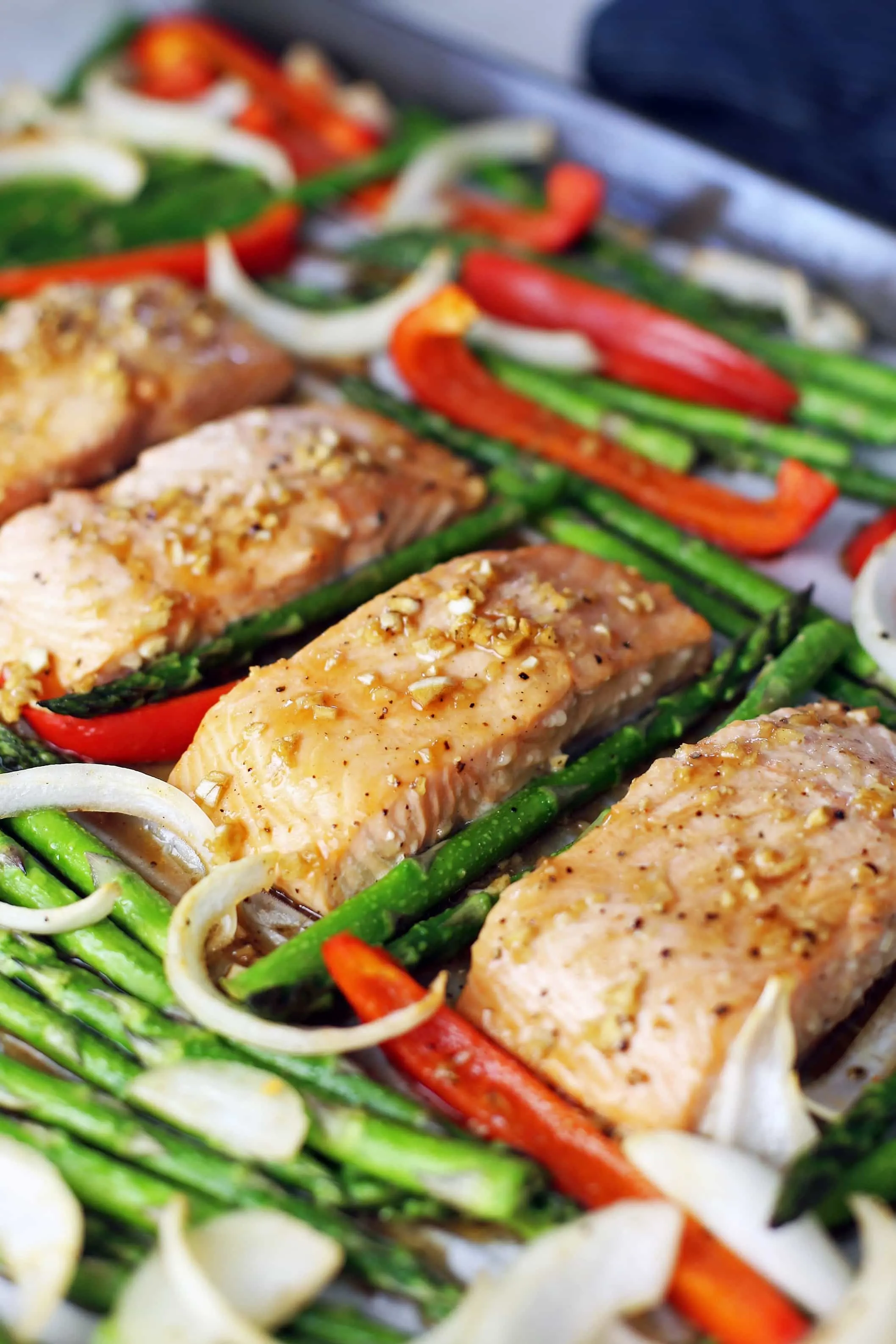 Four baked marinated salmon fillets in a row with roasted asparagus, bell pepper, and onions surrounding them on a large baking sheet.