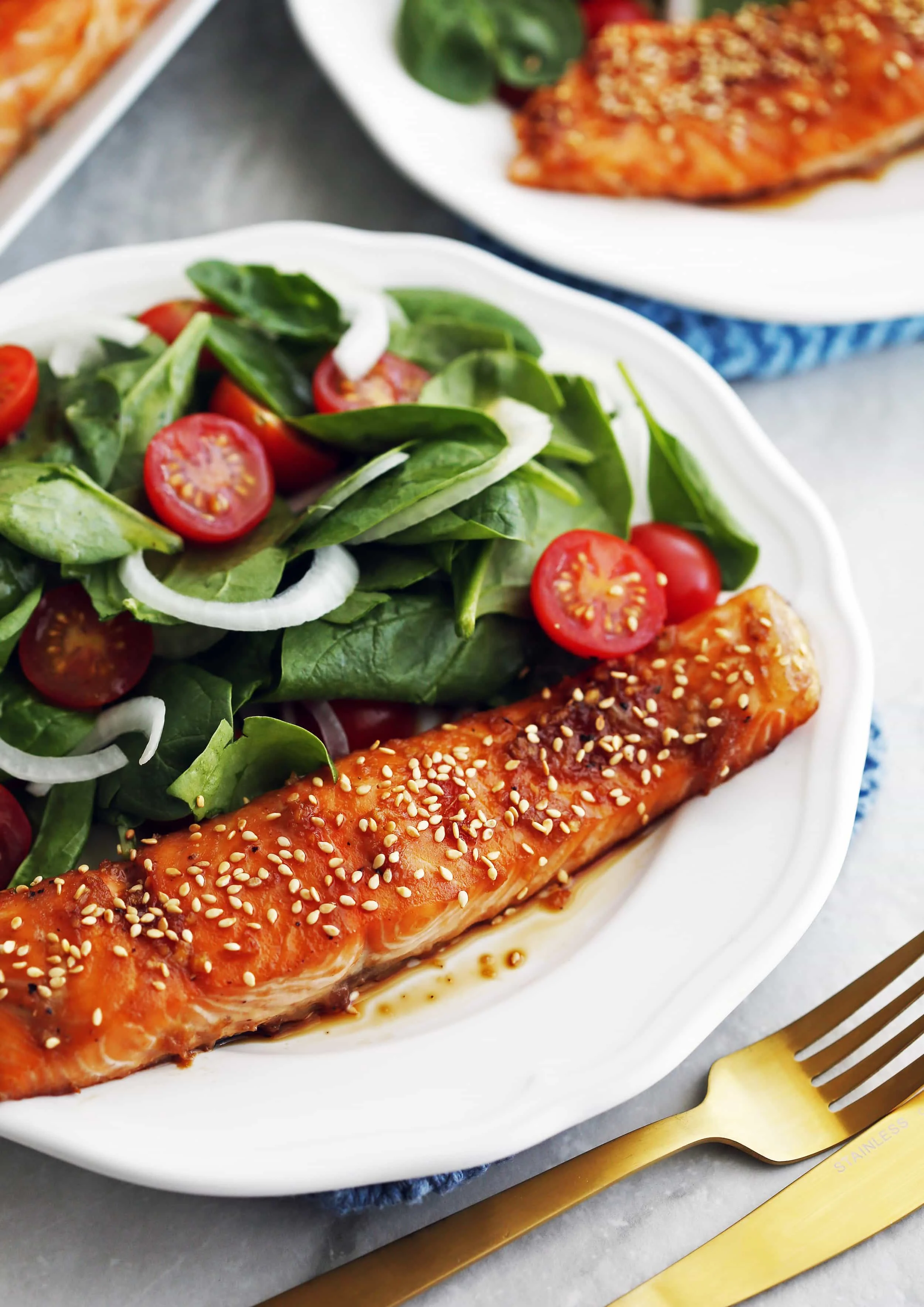 Maple-Soy Baked Salmon with green spinach salad on a white plate with a fork and knife beside it.