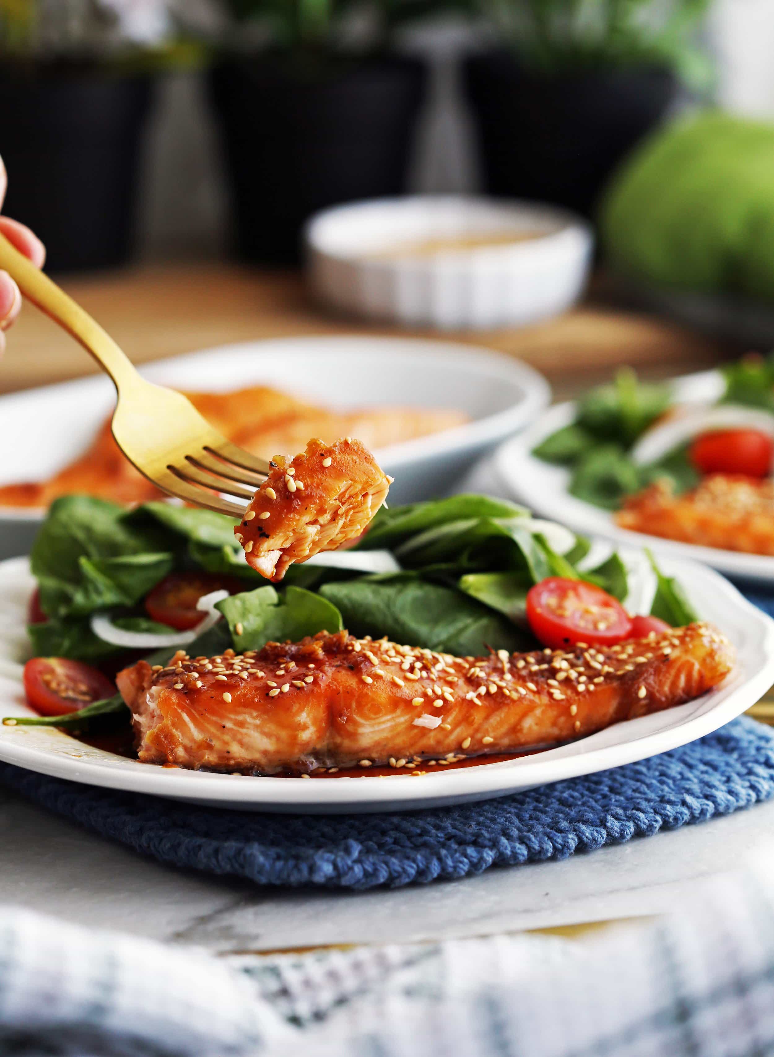 A piece of soy-maple salmon poked with a fork above a top of a plate of a baked salmon with spinach salad.