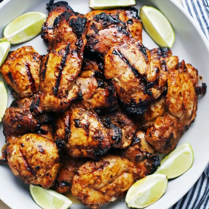 Twelve grilled chili lime chicken thighs on a large grey platter surrounded by lime wedges.