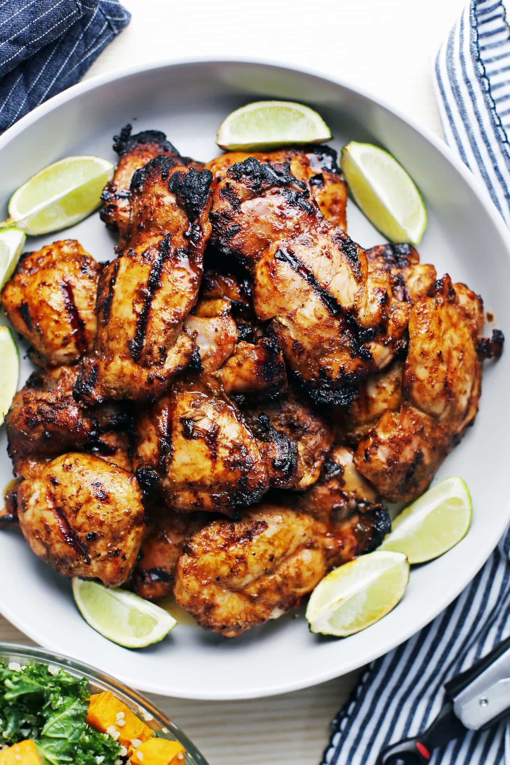 Twelve grilled chili lime chicken thighs on a large grey platter surrounded by lime wedges.