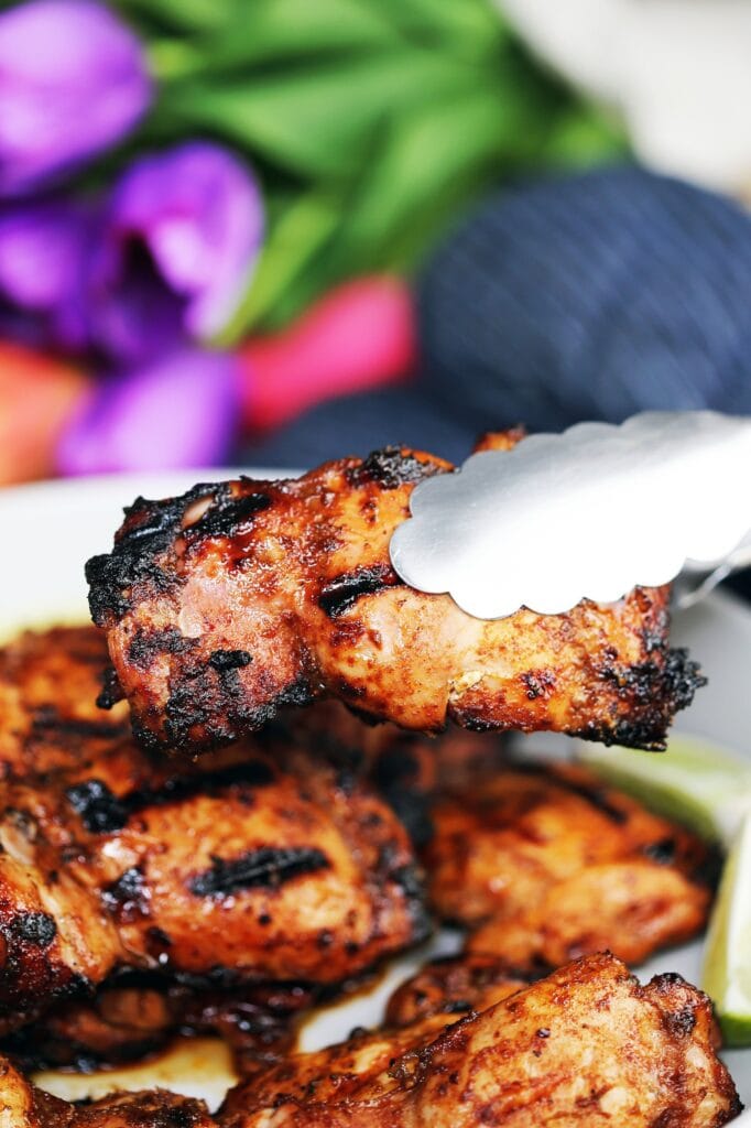 A closeup of a grilled chili lime chicken thigh held by a metal tong over a platter of more grilled chicken thighs.