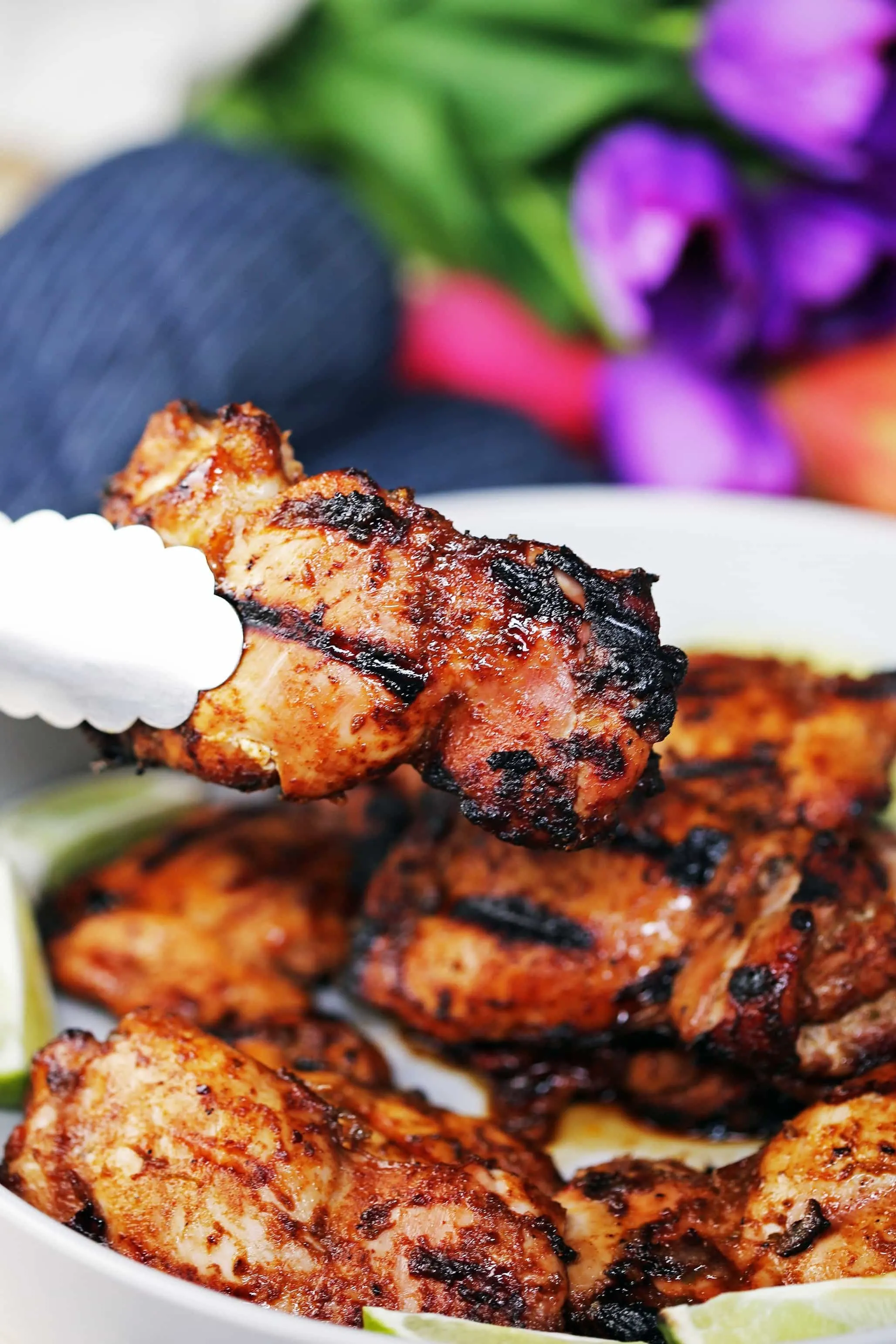 A metal tong holding up a grilled chili lime chicken thigh over a platter of more grilled chicken.