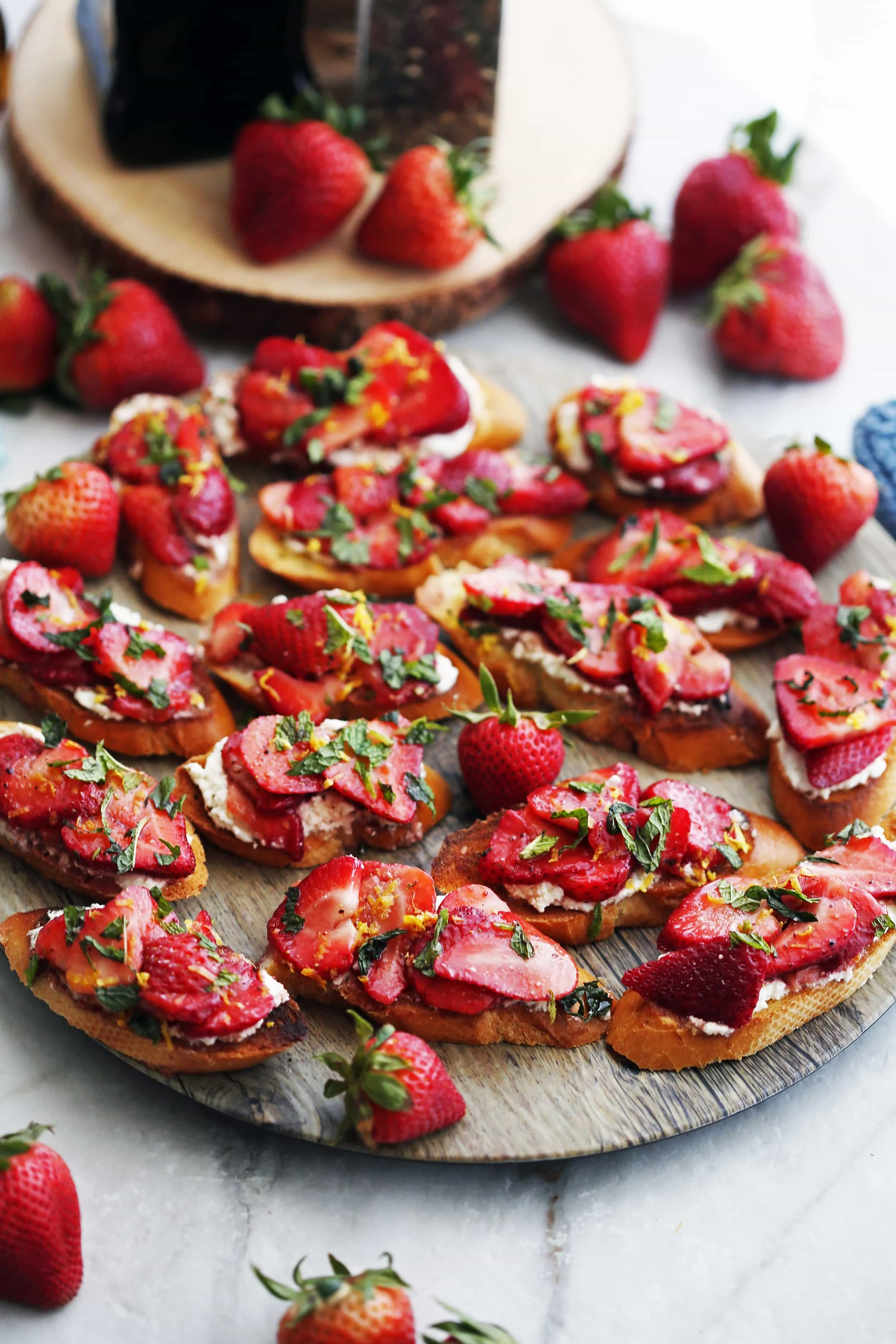 A wooden platter containing fifteen Balsamic Strawberry Ricotta Crostini that's garnished with fresh lemon zest and mint.