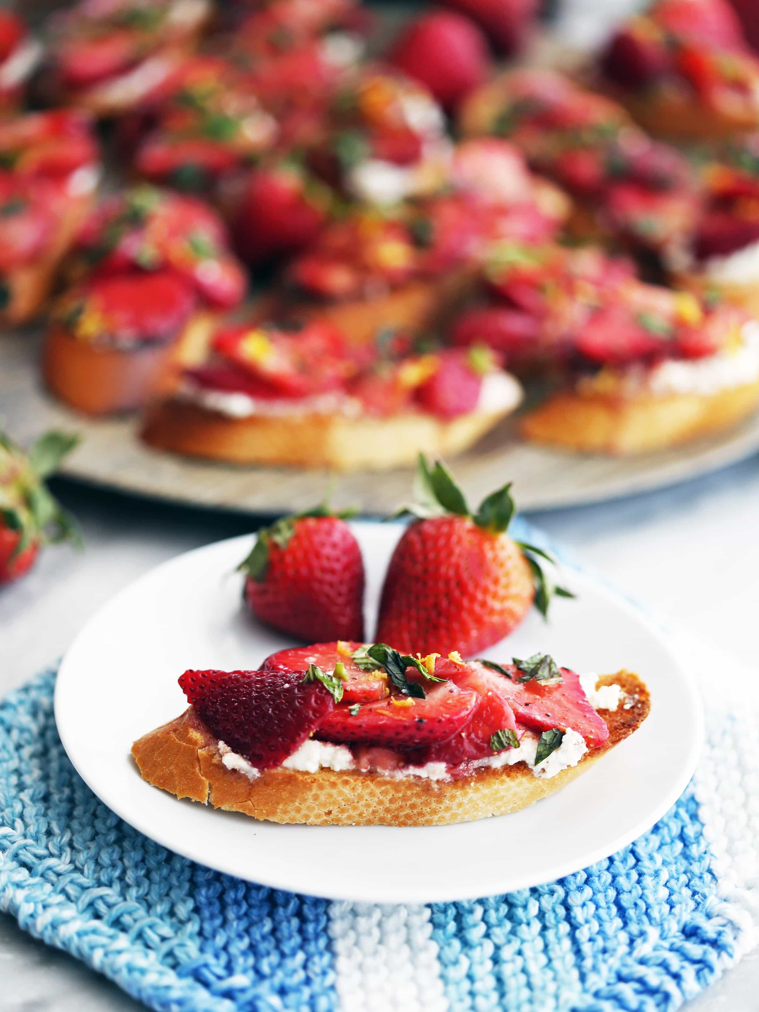 A Balsamic Strawberry Ricotta Crostino on a white plate with two fresh strawberries. More crostini on a platter behind it.