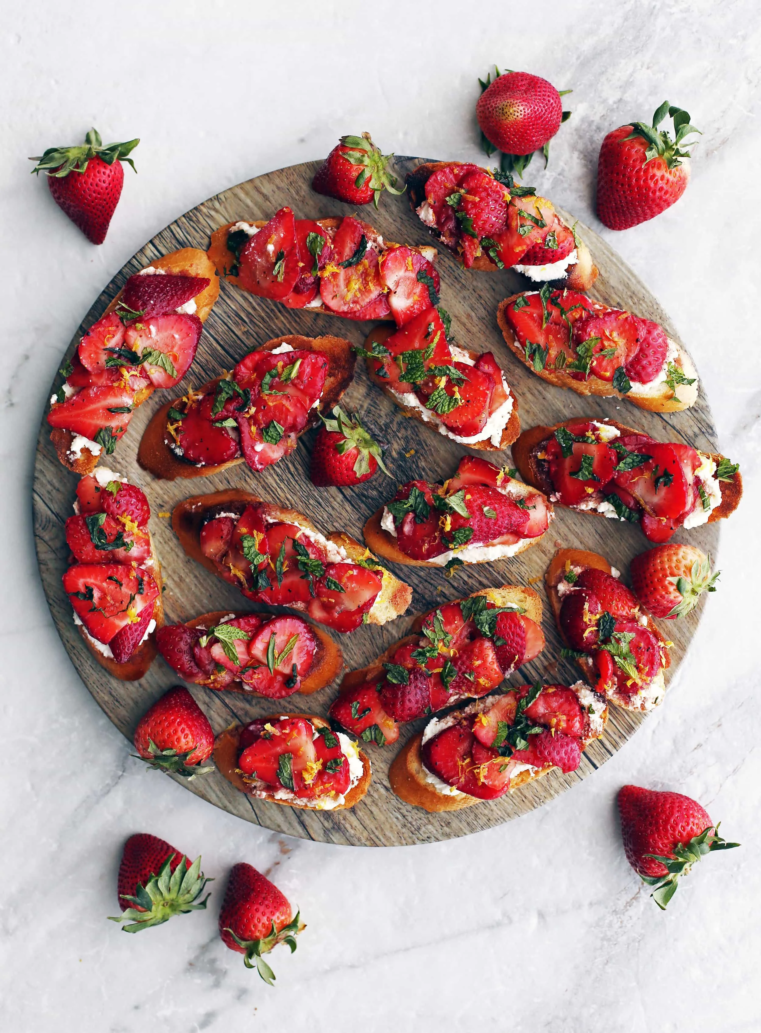 Overhead view of Balsamic Strawberry Ricotta Crostini garnished with mint and lemon zest on a large round platter.