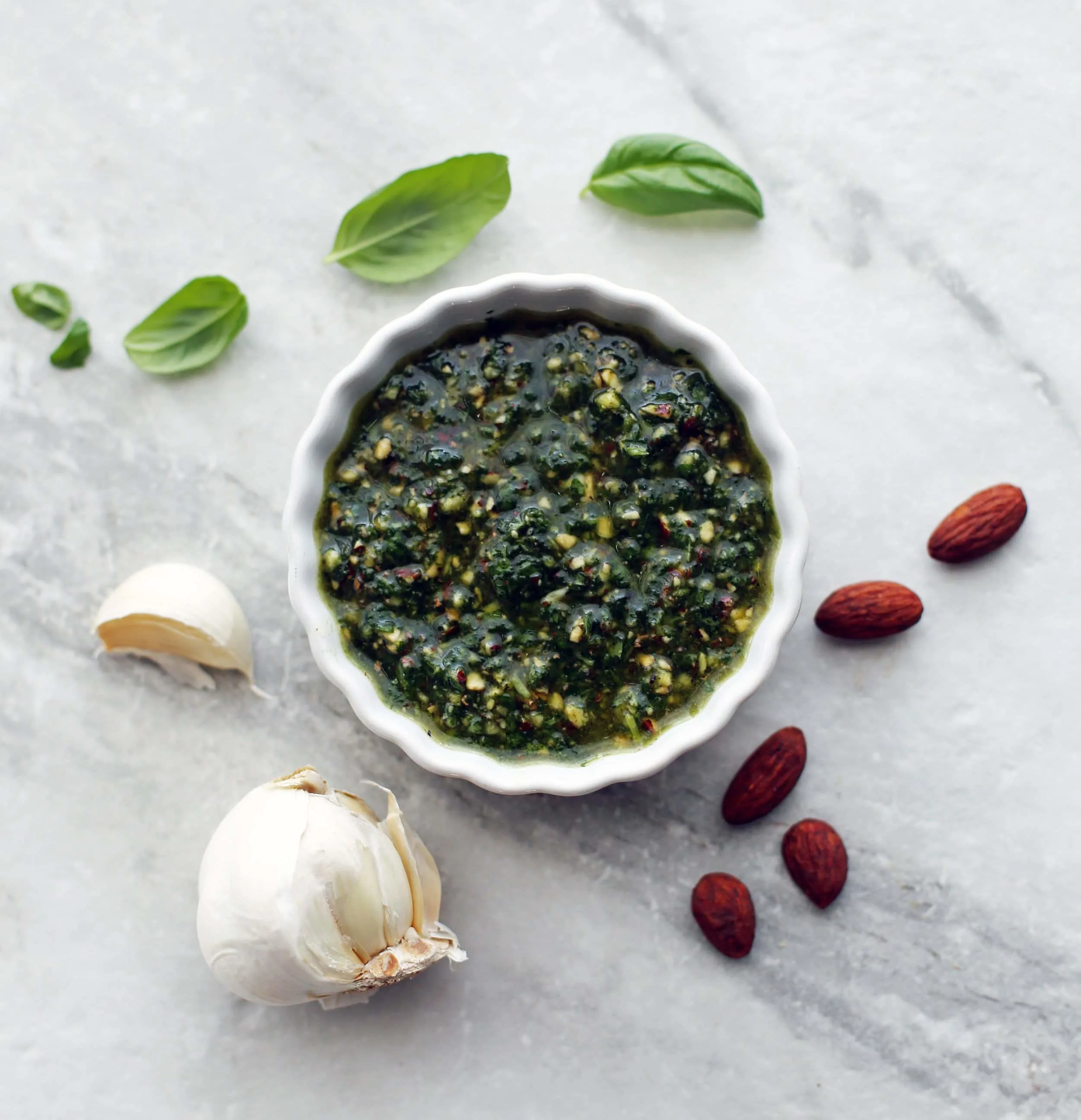 Dark green basil almond pesto in a small white bowl with basil leaves, almonds, and garlic around it.