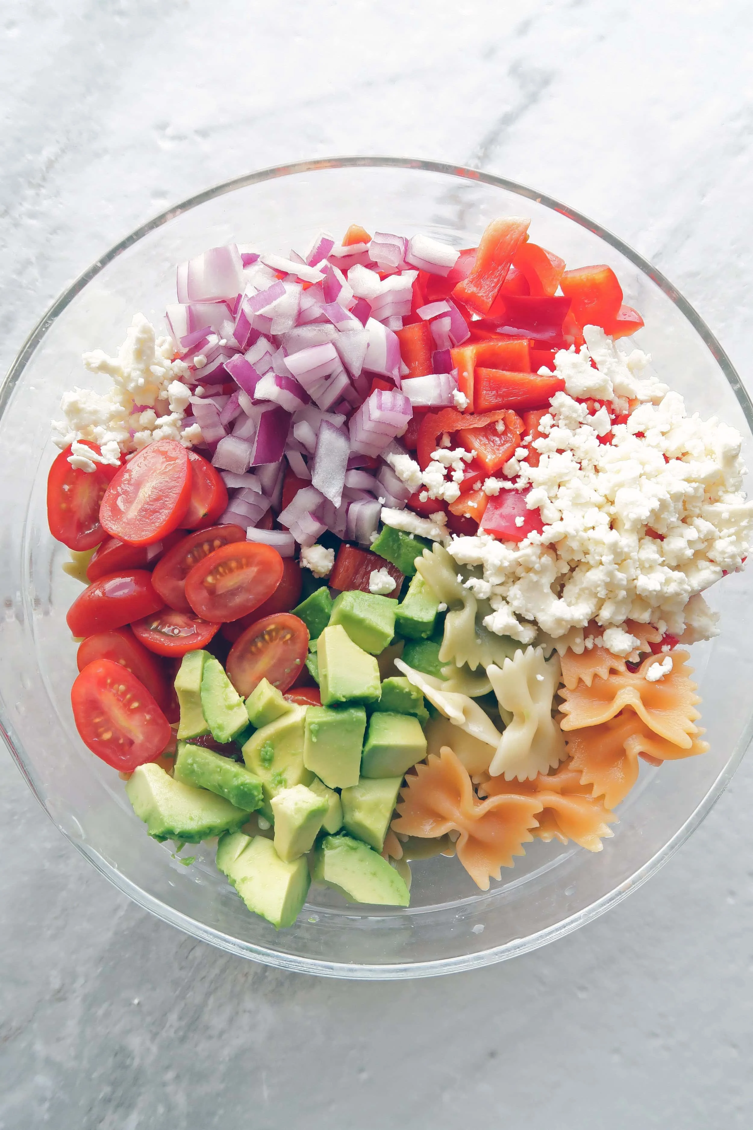 Chopped avocado, tomatoes, red onions, bell pepper, feta, and farfalle pasta in a large bowl.