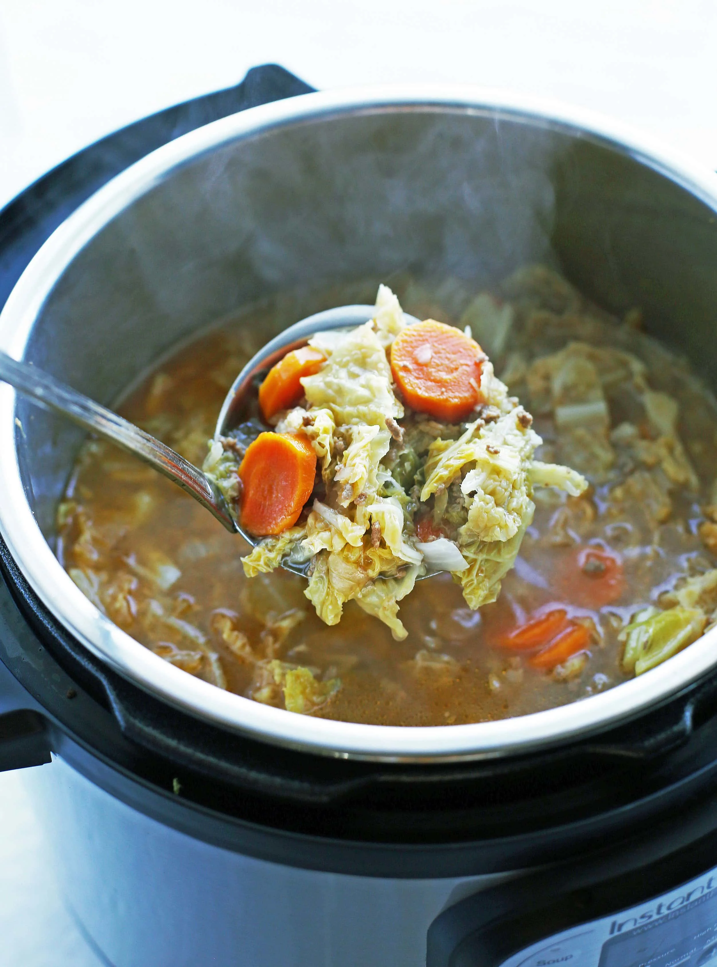 A metal ladle holding a big scoop of Beef and Cabbage Soup over an Instant Pot.