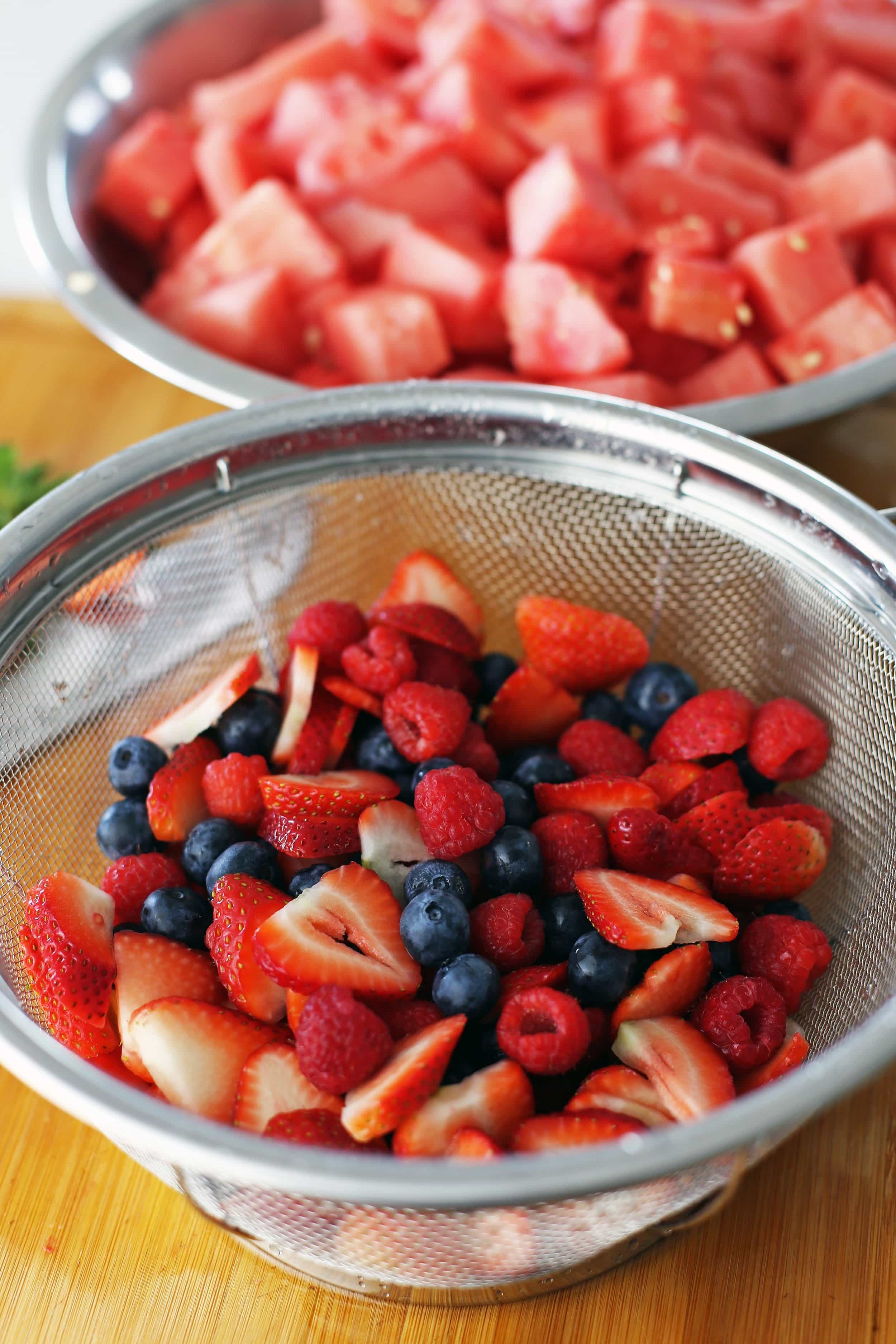Rinsed sliced strawberries, blueberries, and raspberries in a metal stainer with a bowl of watermelon pieces behind it.