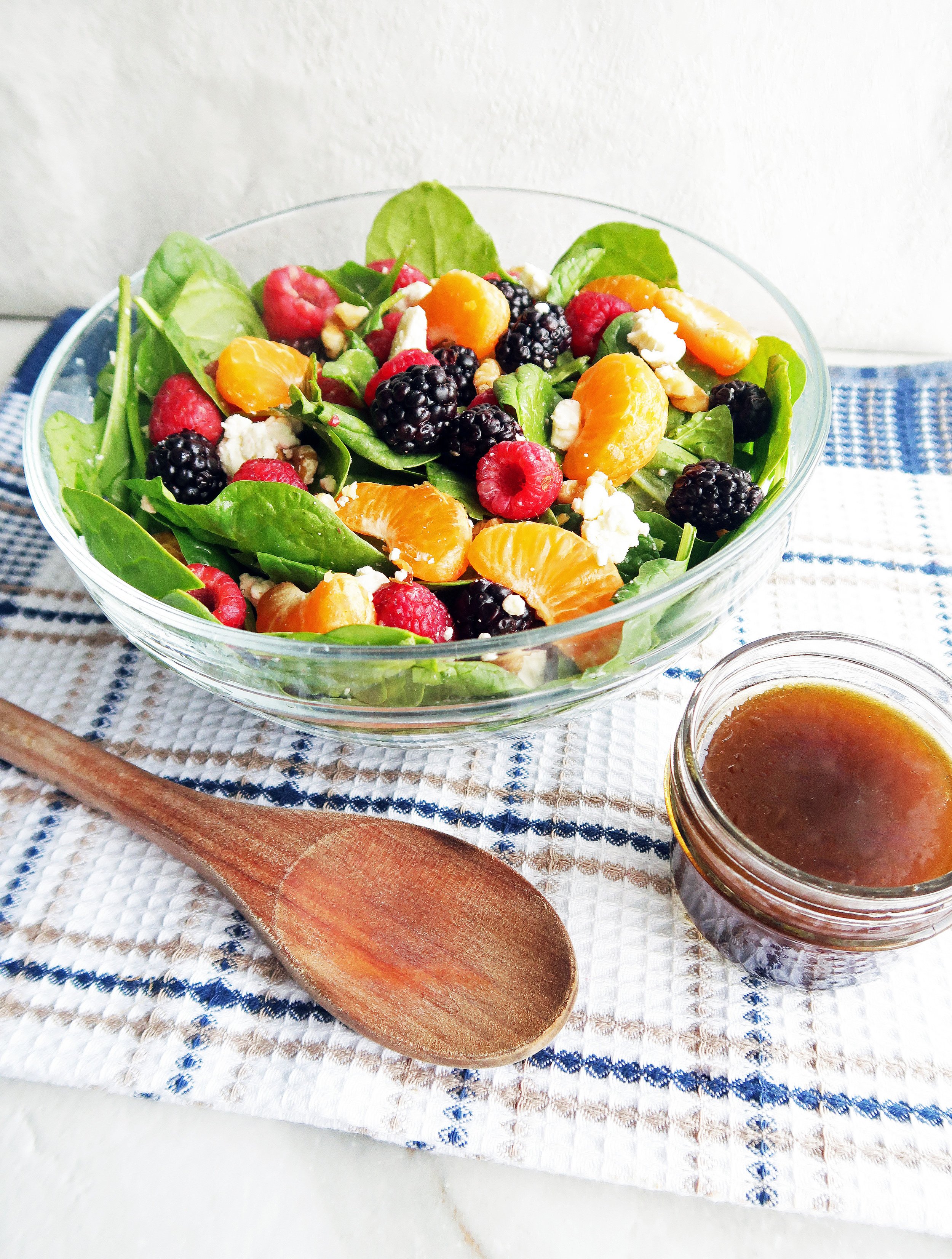 A glass bowl of Berry Orange Spinach Salad tossed in Citrus Balsamic Vinaigrette with more dressing in a small jar and a wooden spoon to the side.