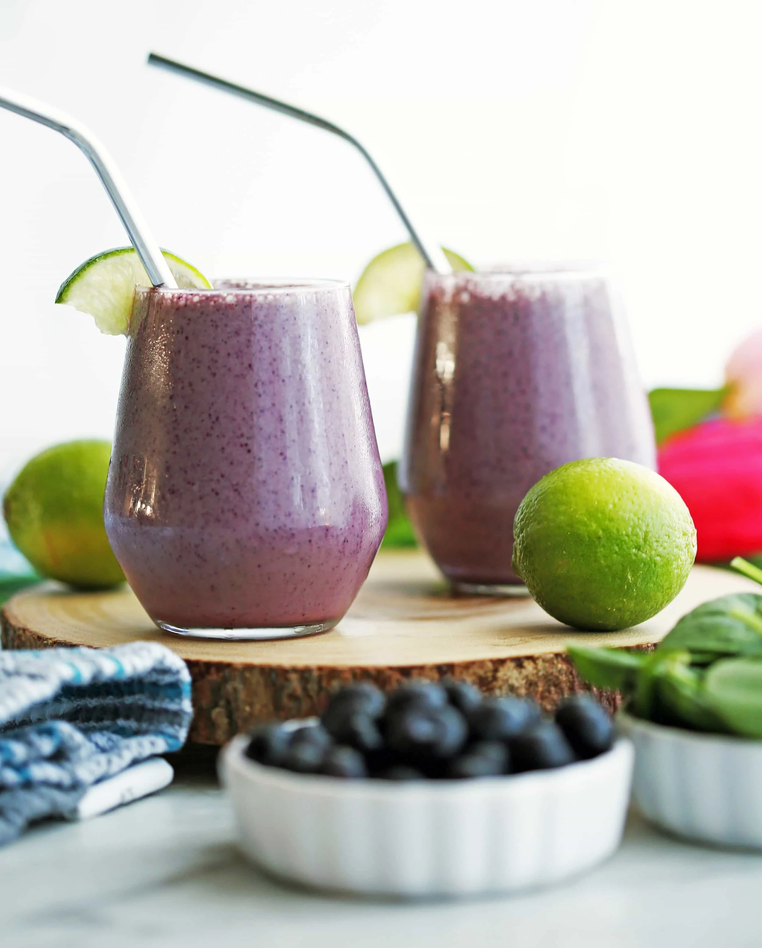 Two Blueberry Lime Yogurt Smoothies with lime wedges and straws on a wooden board.