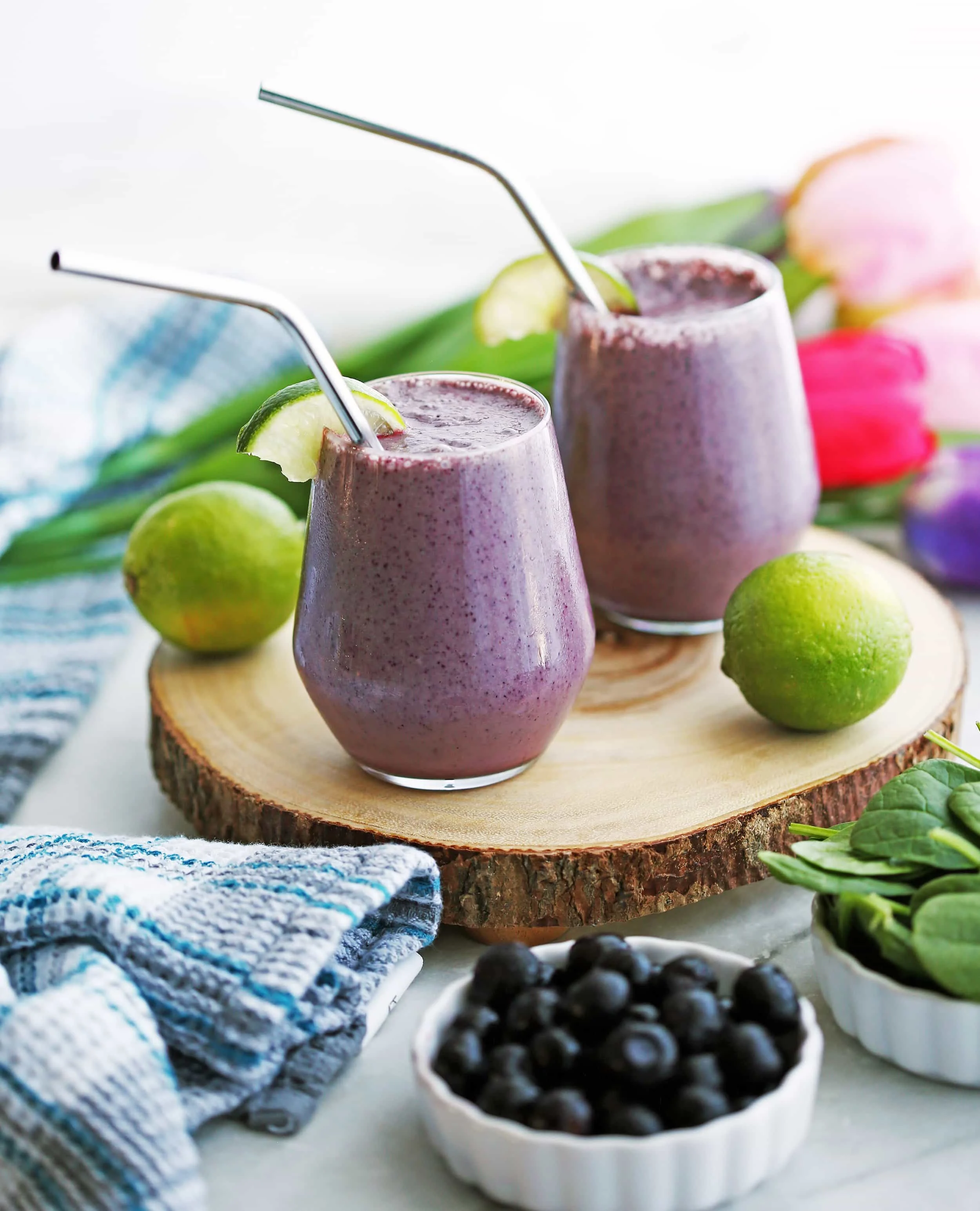Two Blueberry Lime Yogurt Smoothies on a round wooden board, surrounded by limes, blueberries, and spinach.